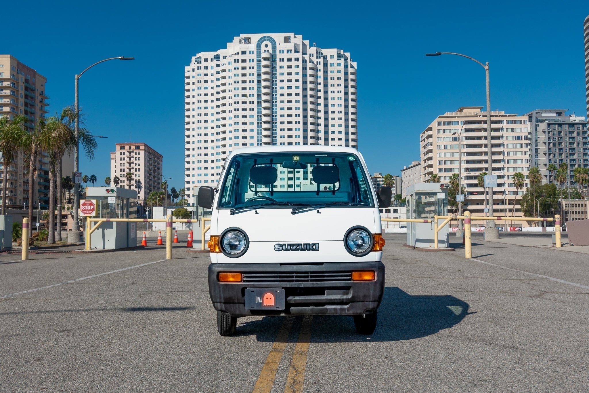 A white Suzuki van is parked on a street with tall buildings and palm trees in the background.