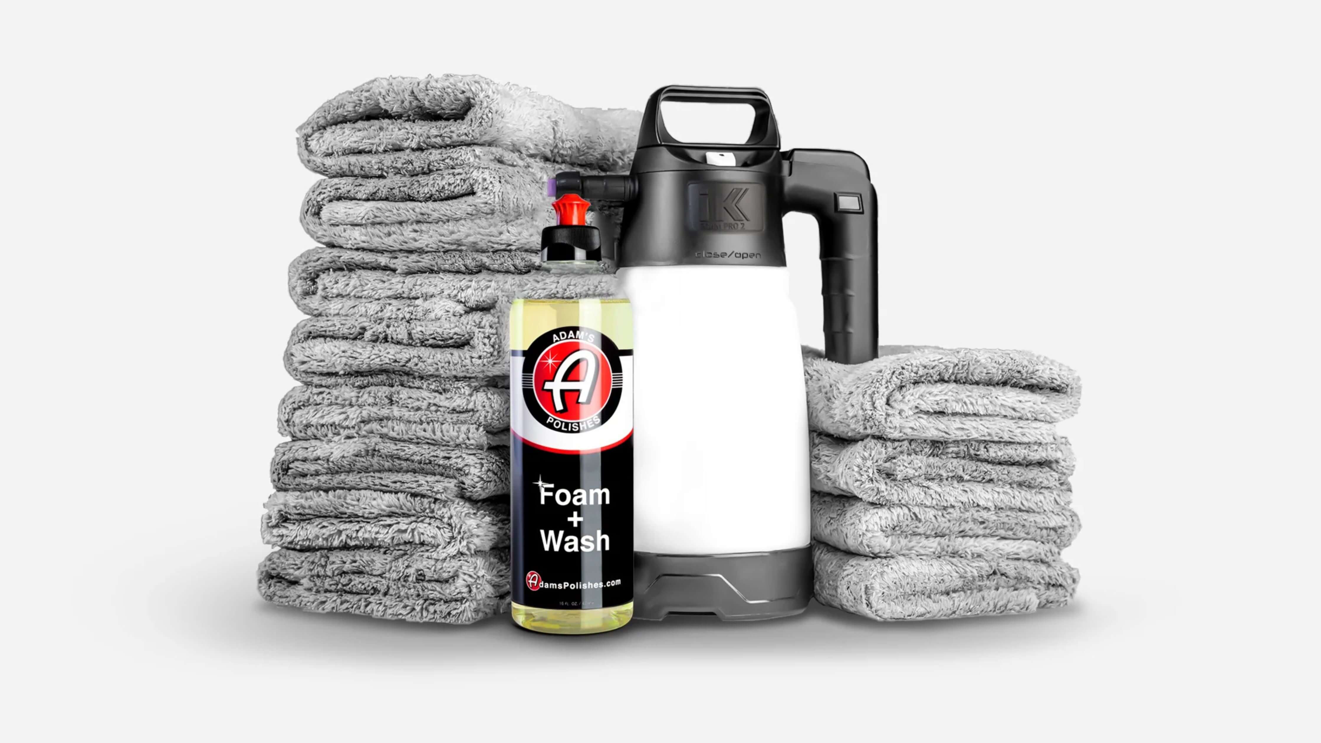 A collection of gray microfiber towels, a bottle of Adam's Polishes Foam + Wash, and a foam sprayer are arranged on a white background.