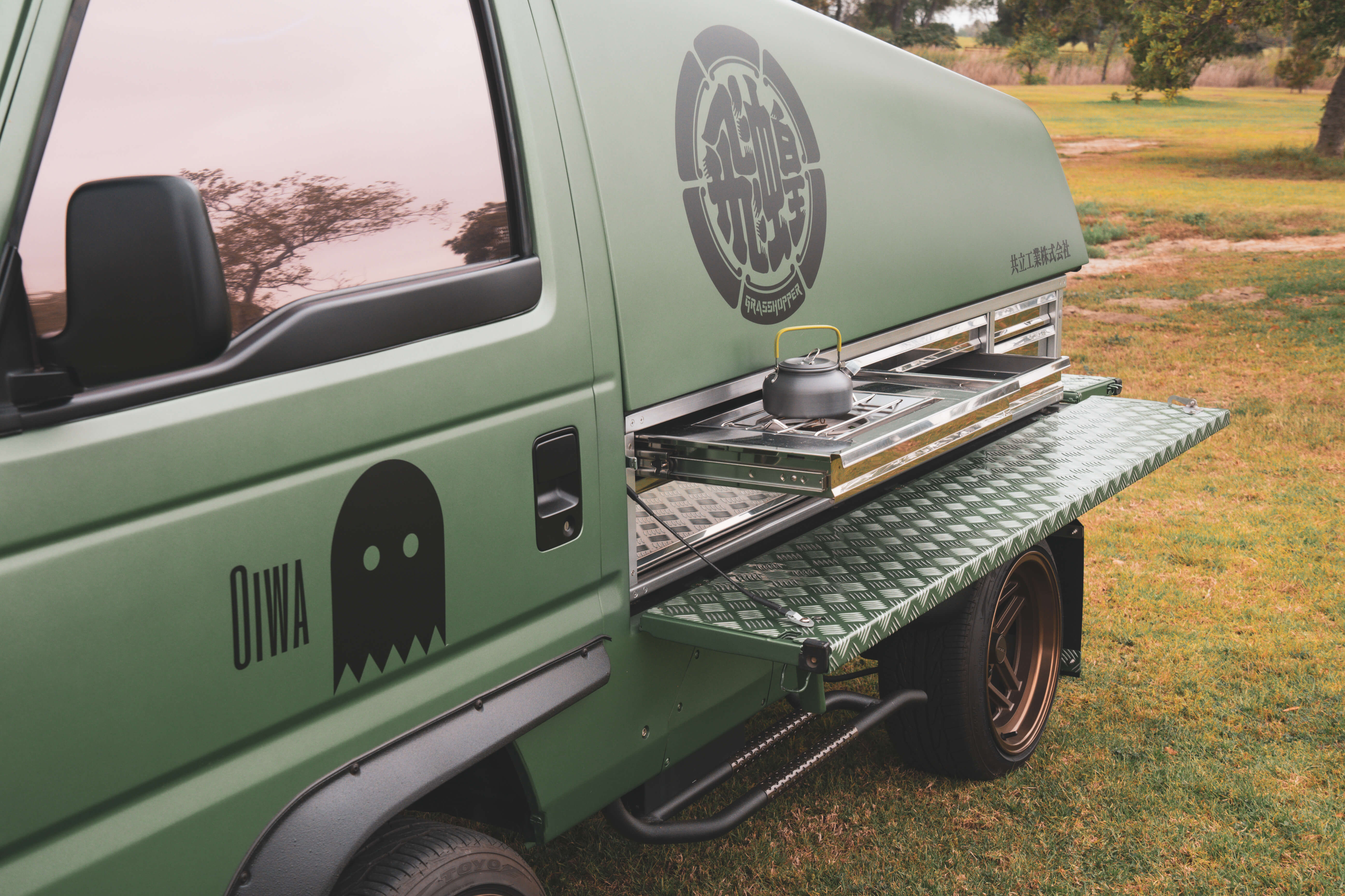 Custom Grasshopper-branded Kei Truck by Oiwa Garage featuring a sleek matte green exterior, integrated pull-out camping kitchen, kettle on stove, bronze wheels, and checkered drop-down side panel for outdoor adventures.