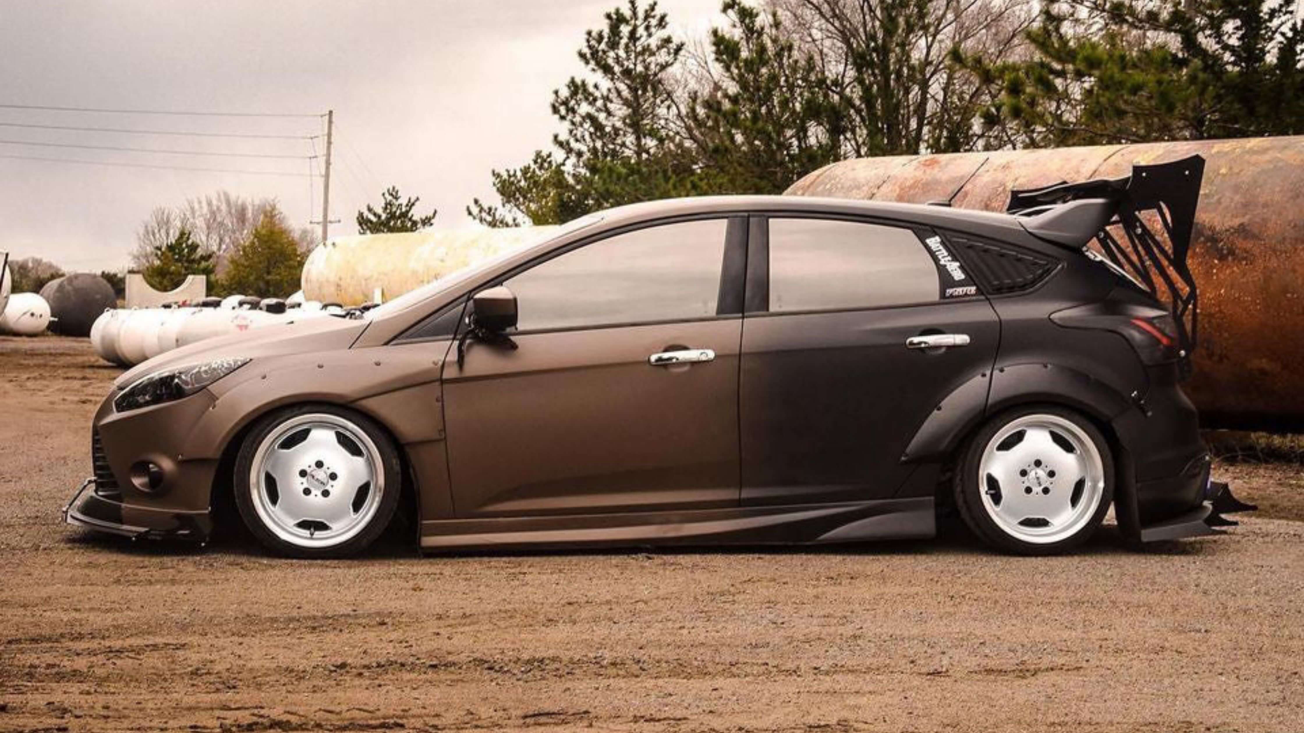 A modified car with a matte finish, large rear wing, and custom wheels on a dirt surface with industrial tanks in the background, showcasing car tint effects.