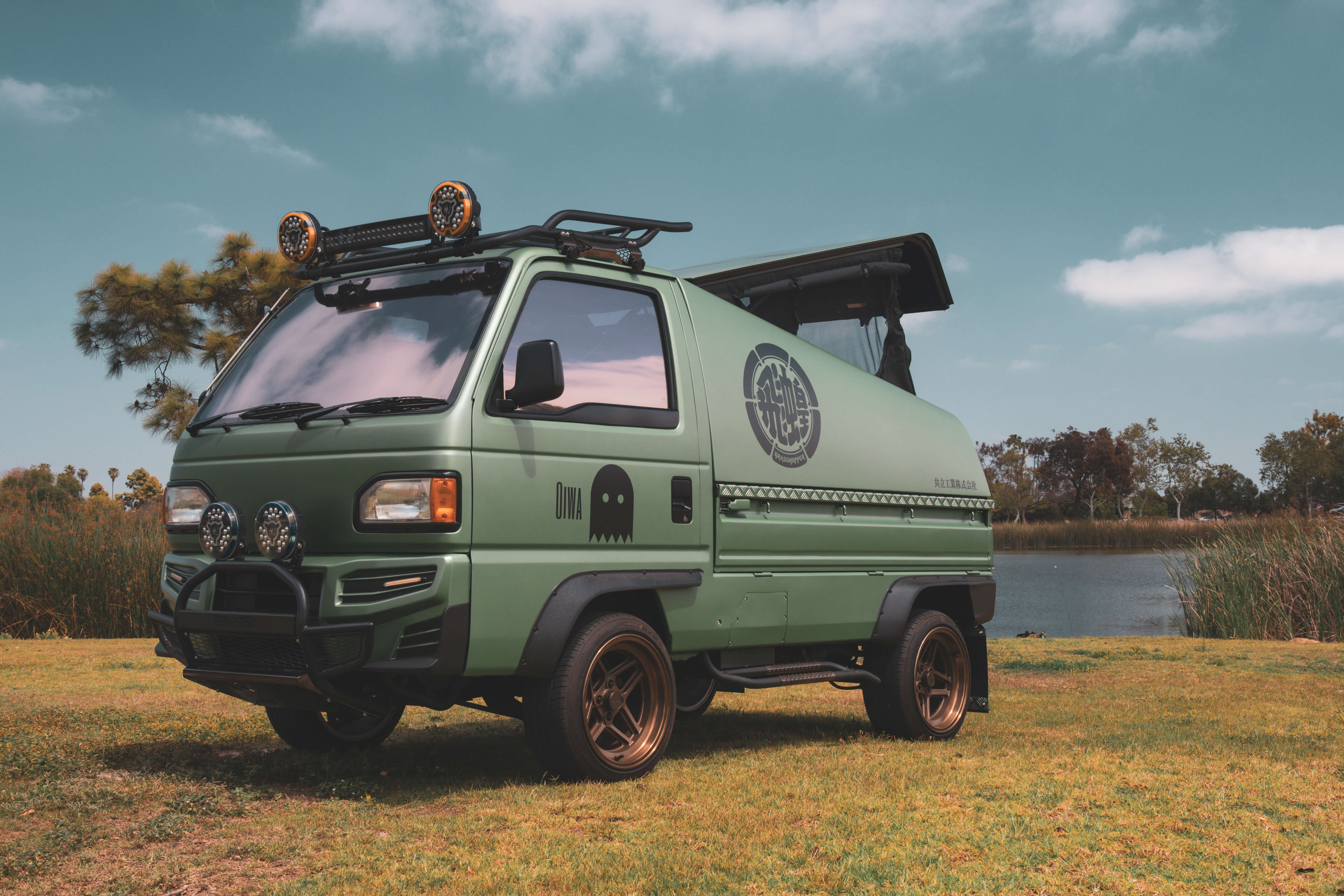 Customized green Honda Acty 4x4 kei truck with roof rack, off-road lights, and popup camper parked outdoors near a scenic lake.