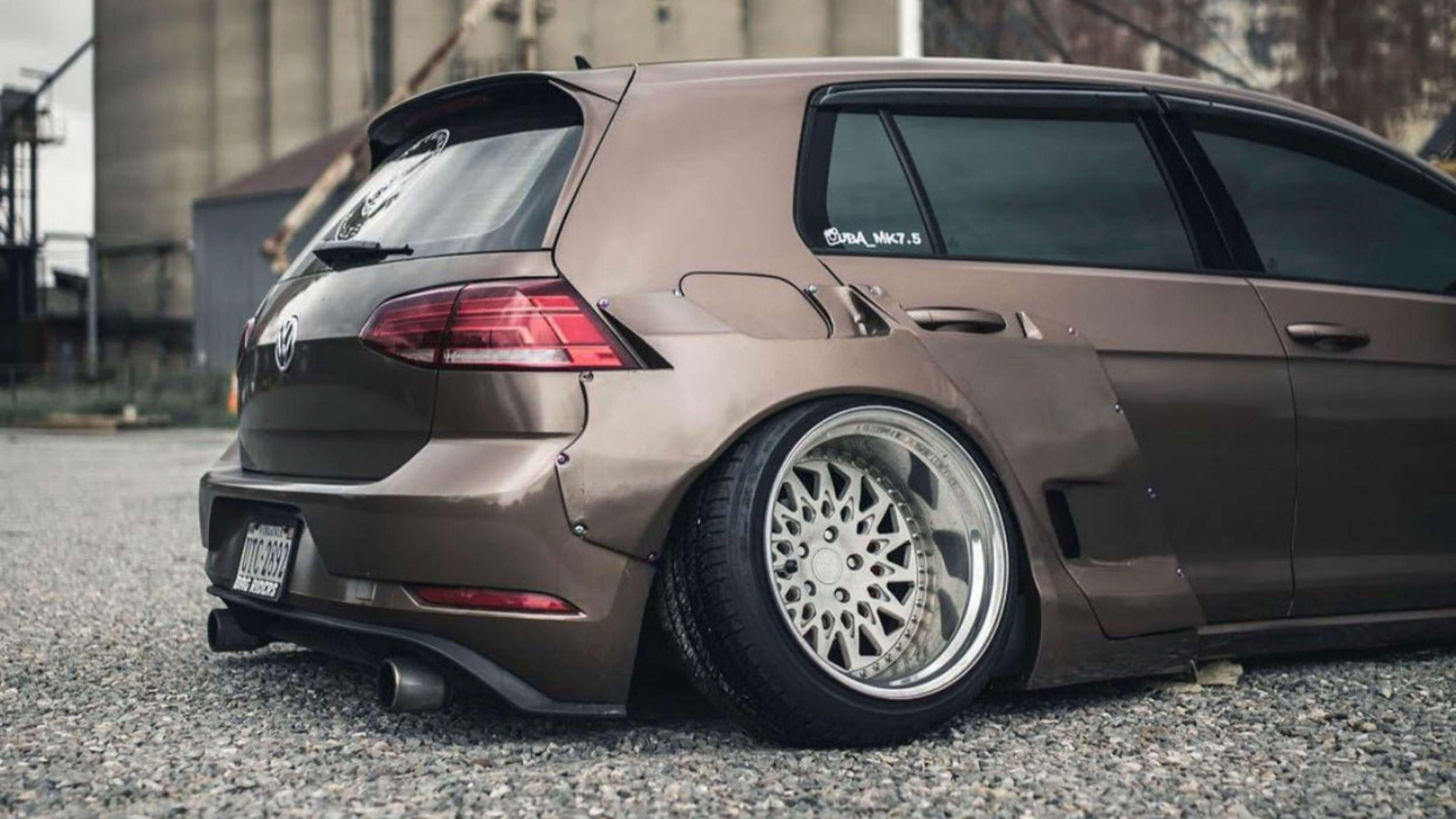 A modified brown Volkswagen hatchback with wide wheels and a lowered stance, parked on a gravel surface.