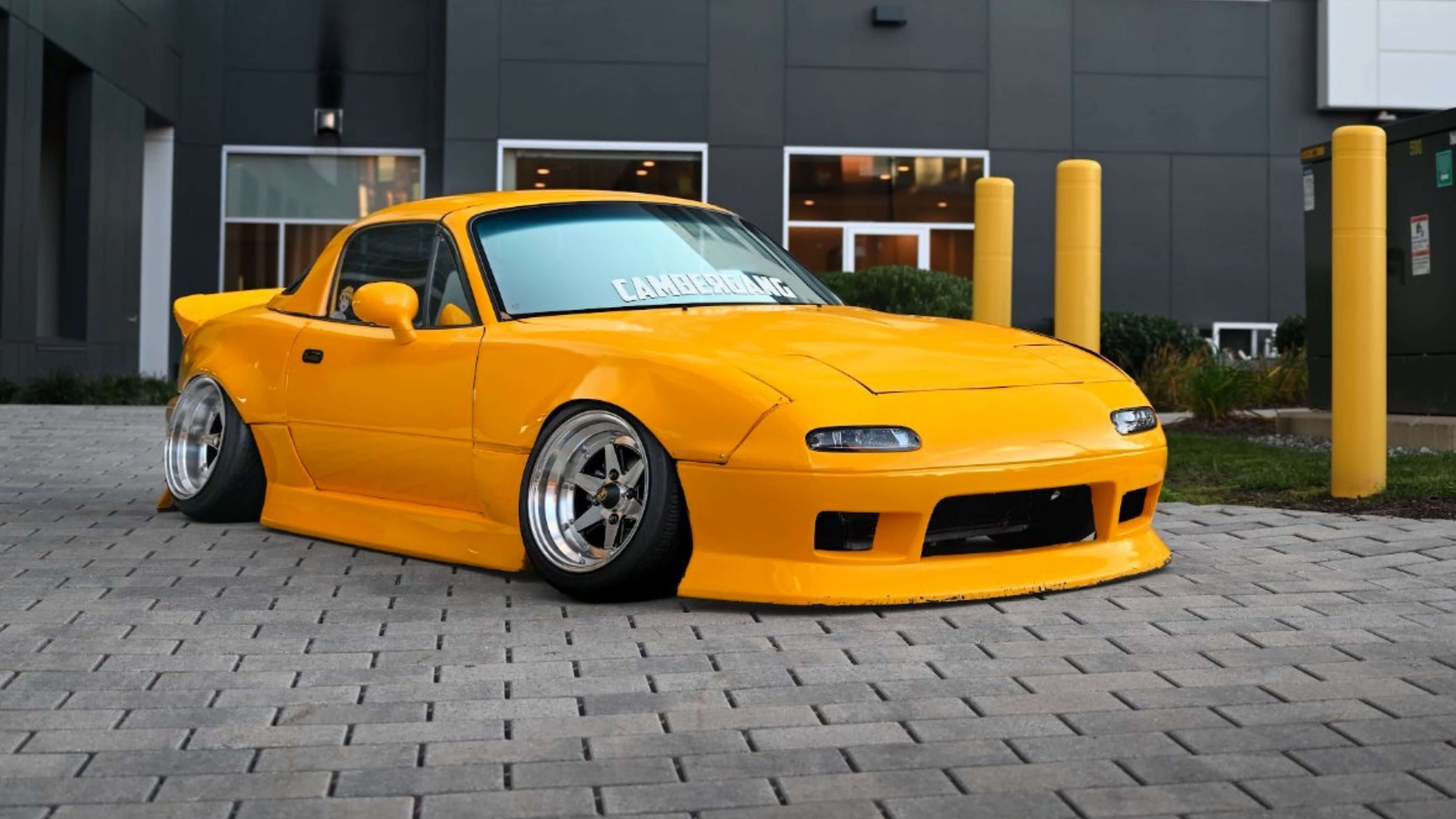 A bright yellow, modified sports car with a low stance and wide wheels parked on a paved area in front of a modern building.