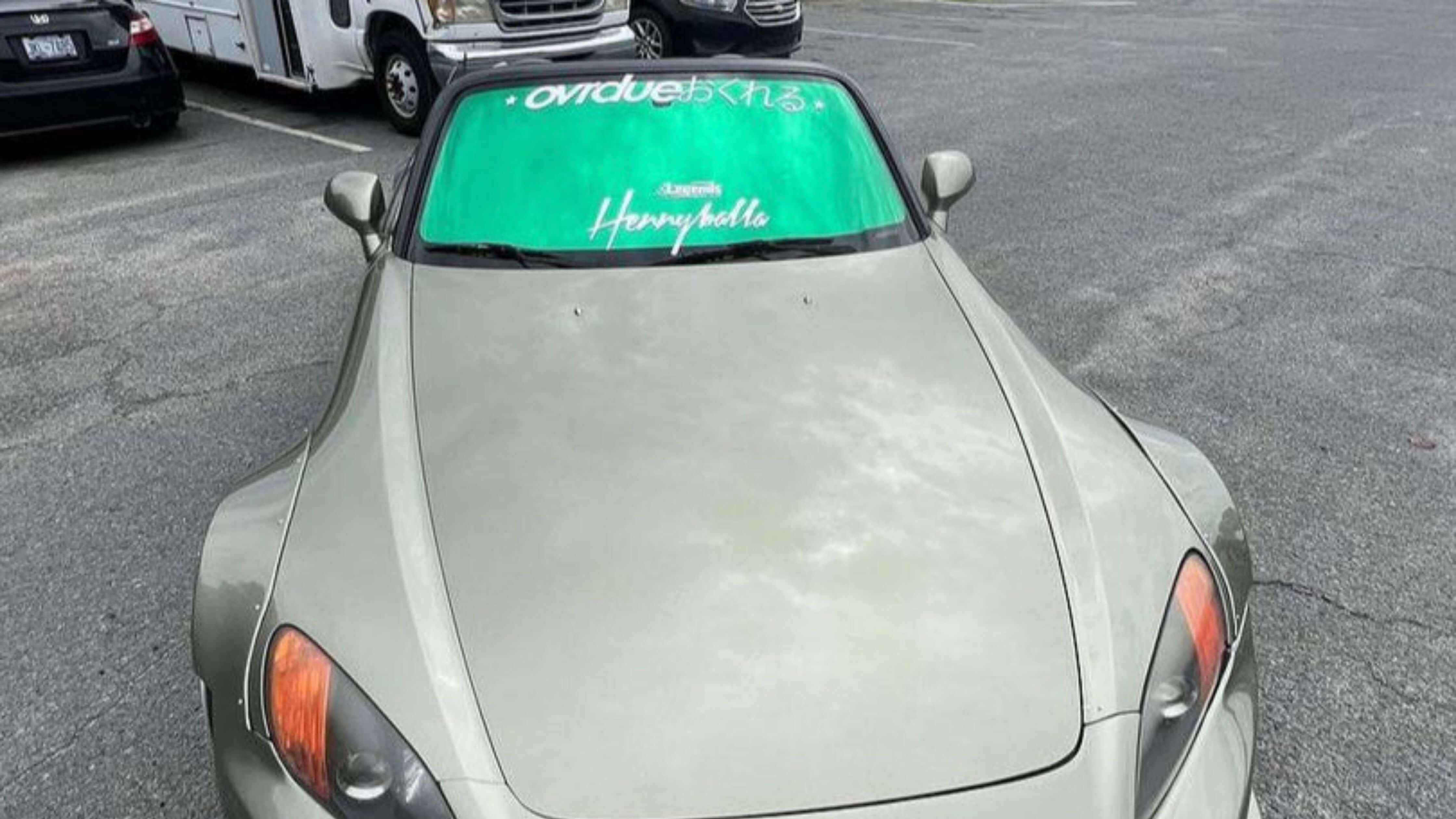 Light gray car with green windshield sunshade reading 'overdue' and 'Hennyholla' in parking lot.