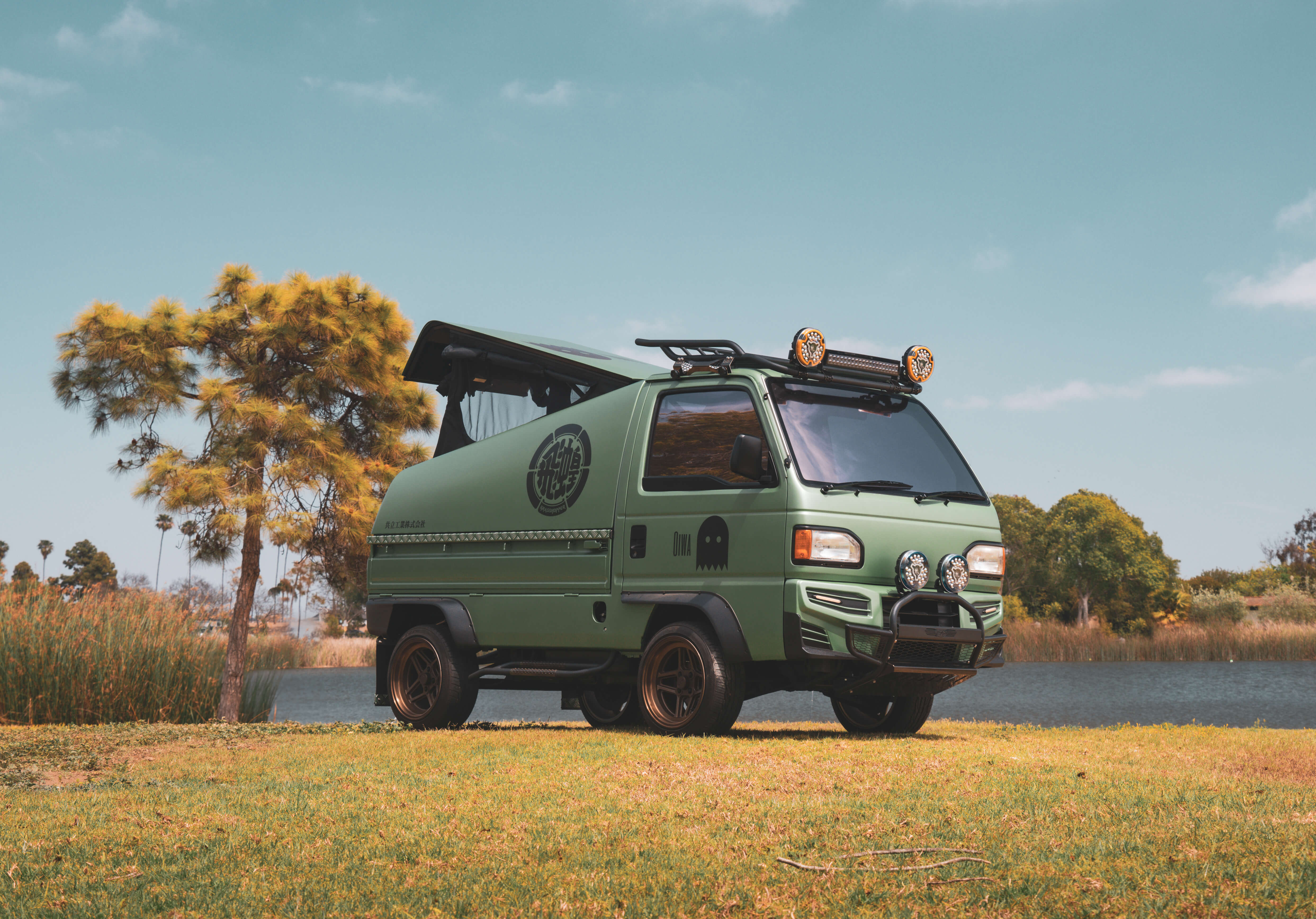 Green Honda Acty Grasshopper kei truck camper with pop-up metal camper shell, lifted suspension, roof rack, and off-road lighting parked in scenic outdoor setting.