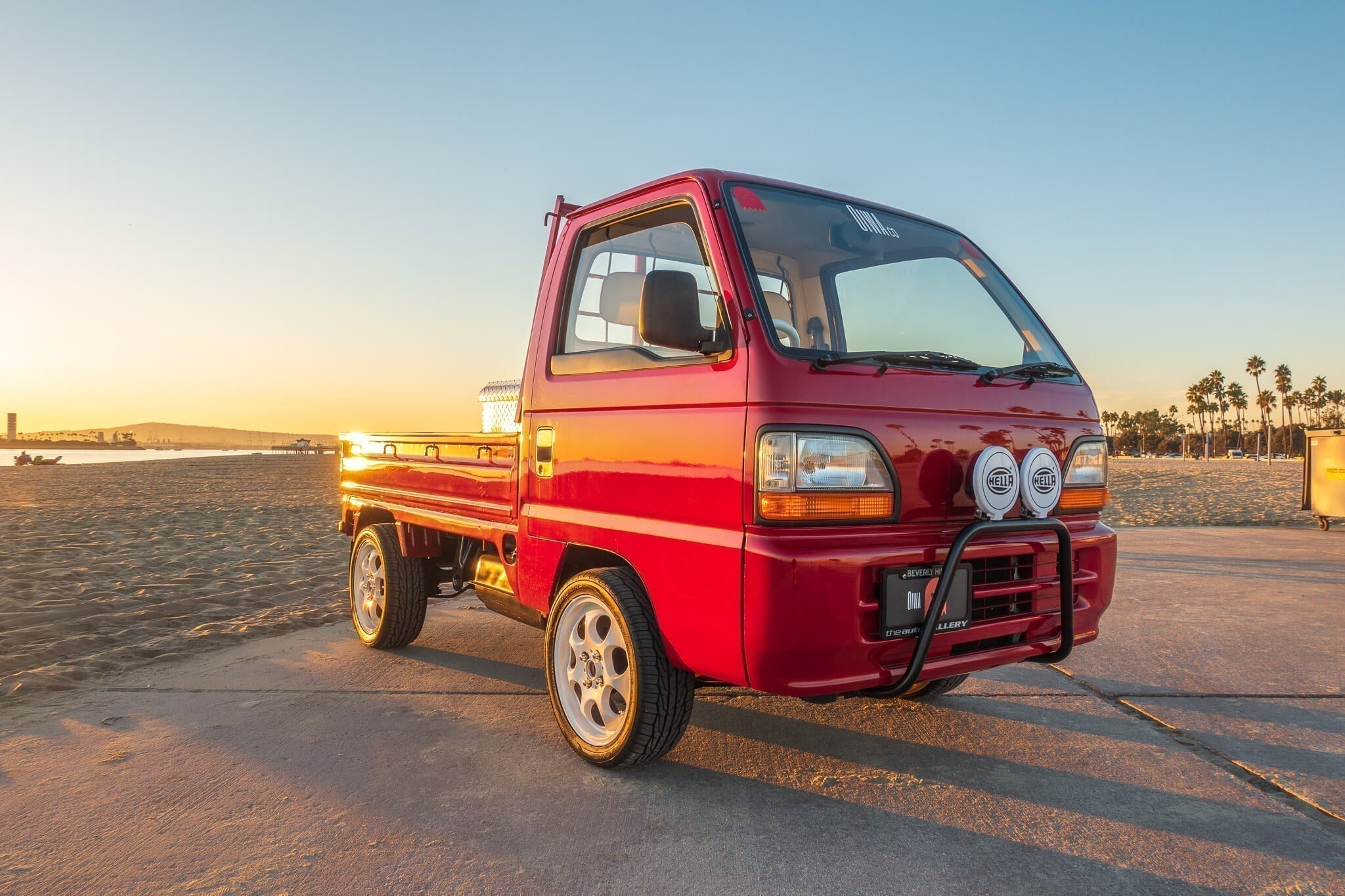 Red Honda Acty mini truck parked on a beach at sunset, with palm trees in the background.
