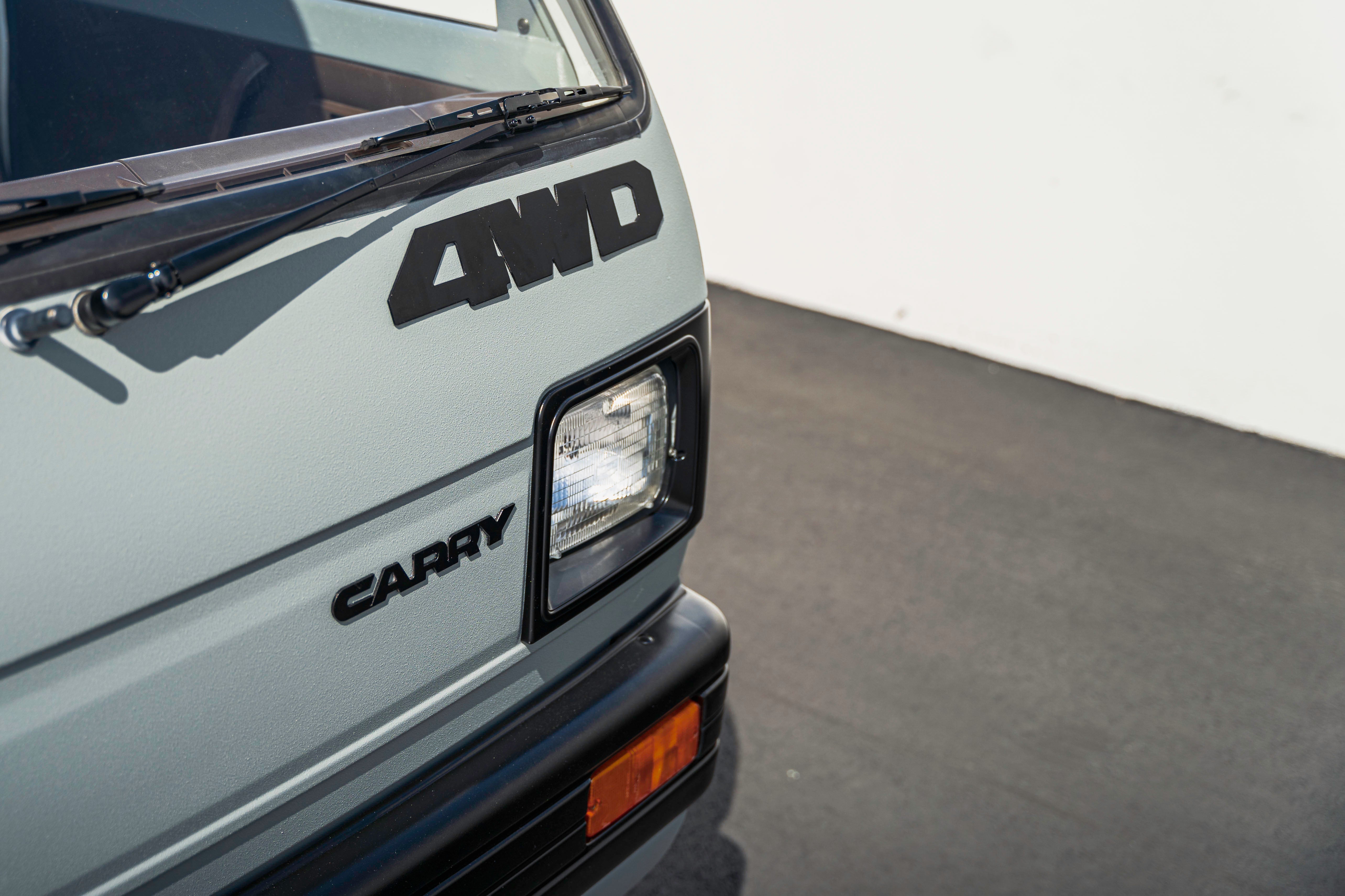 Close-up of the front corner of a vehicle with "4WD" and "CARRY" badges, showing a headlight and windshield wiper.