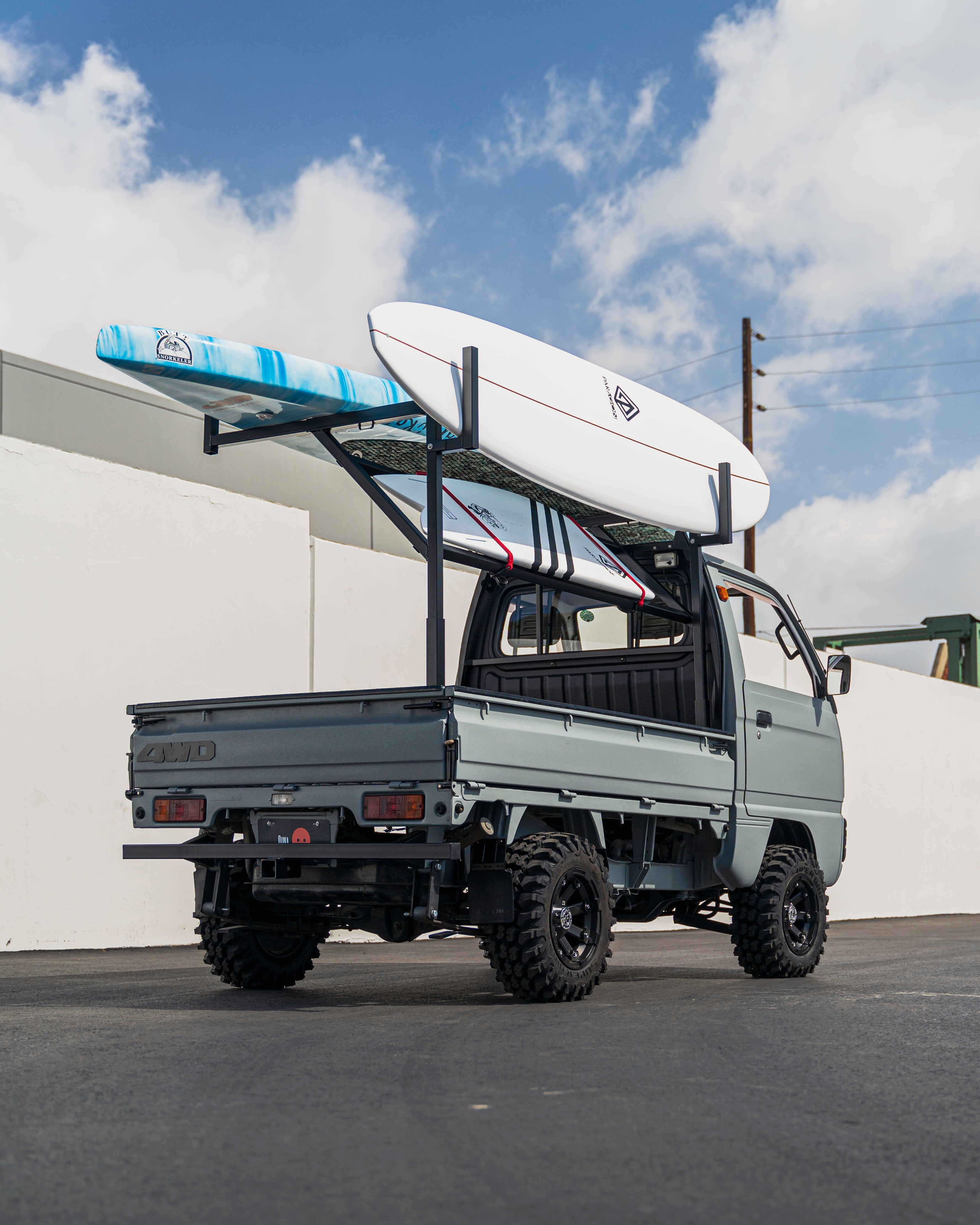 A small, rugged 4WD truck with surfboards mounted on a rack, parked on a paved surface against a white wall and blue sky.