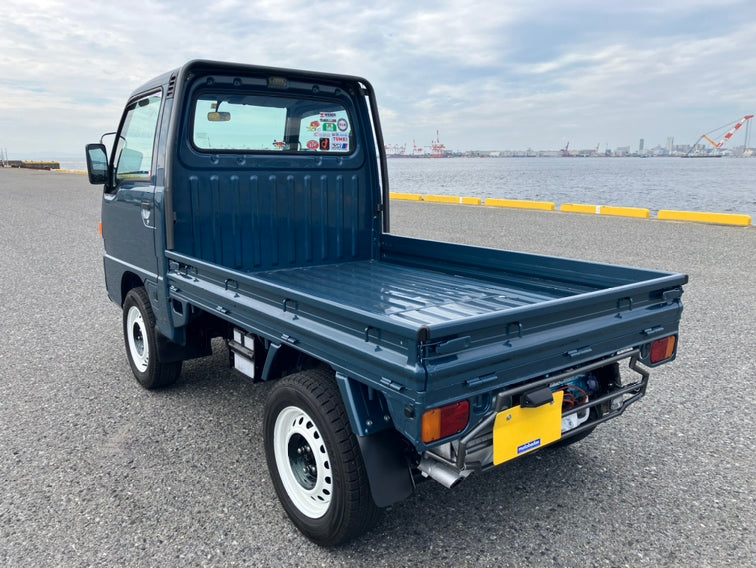 1990 Subaru Sambar parked on a gravel surface near waterfront cranes.