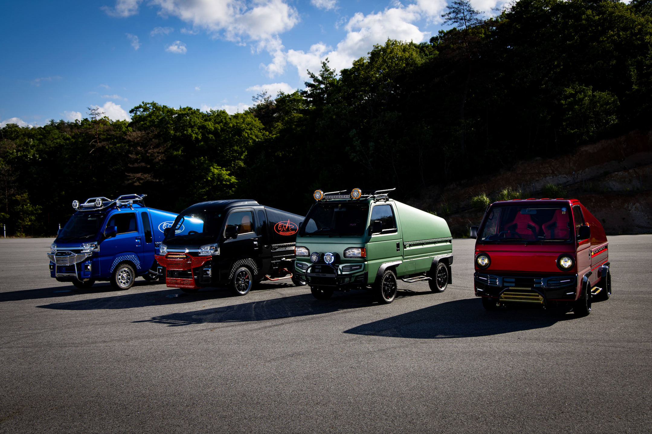 A lineup of colorful modified kei trucks in an outdoor parking lot, showcasing a range of customizations including roof racks, extra lights, and specialty decals, representing Oiwa Garage’s diverse collection of Japanese mini trucks.