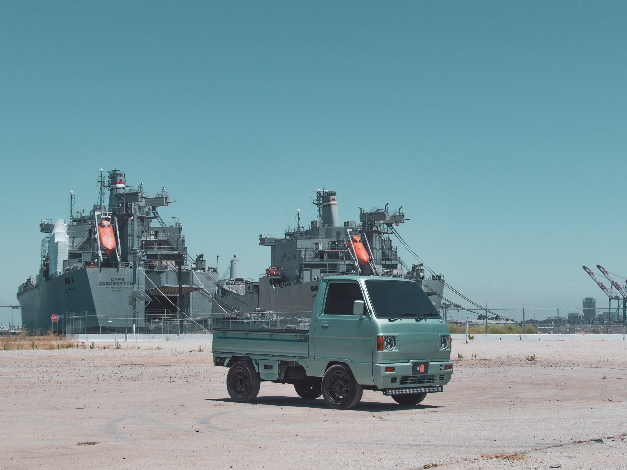 Mint-green 6x6 kei truck for sale, parked on sandy lot with ships and cranes in maritime setting.