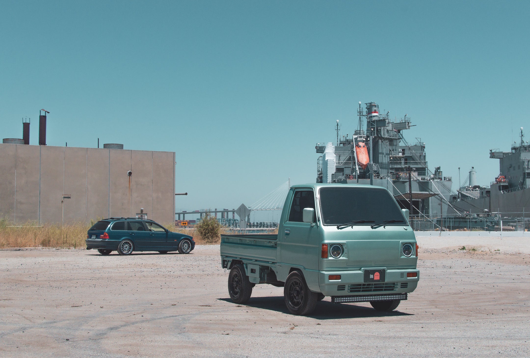 Light green kei truck parked on dusty lot with blue sky and industrial ships, showcasing compact size and utility in a maritime setting.