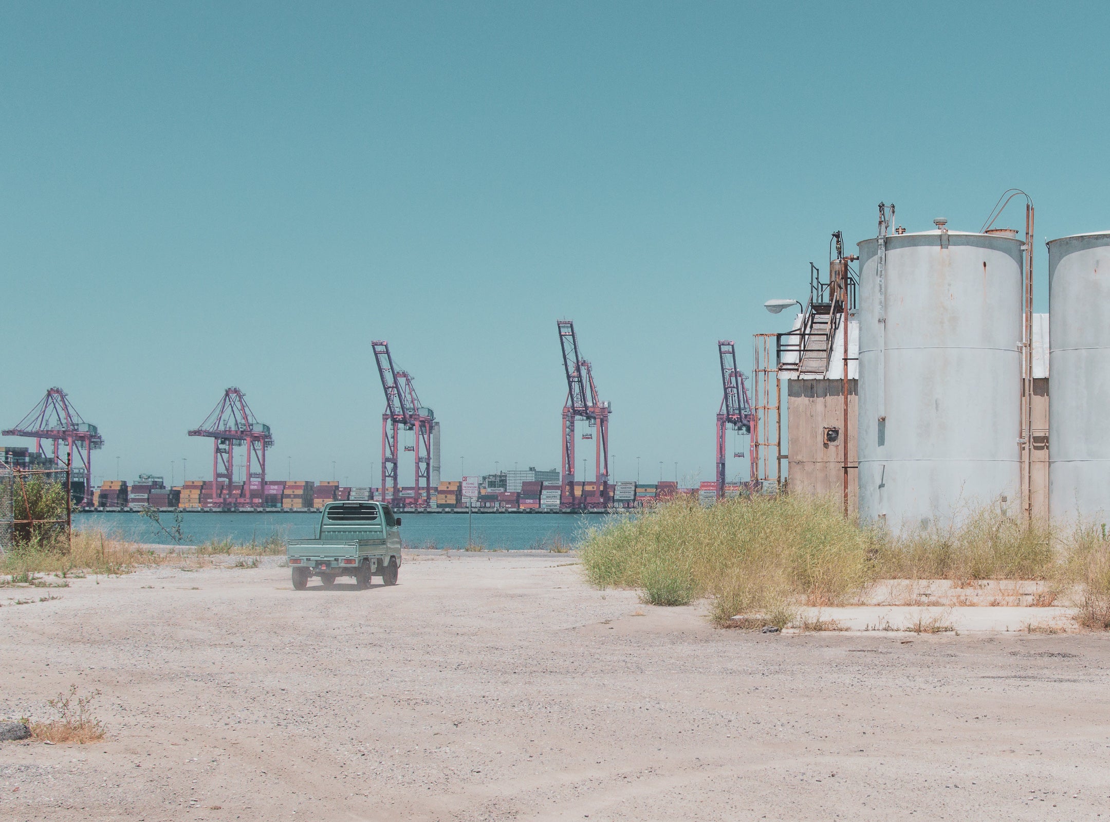 Modded kei truck on gravel path near water, with pink cranes and containers in background.