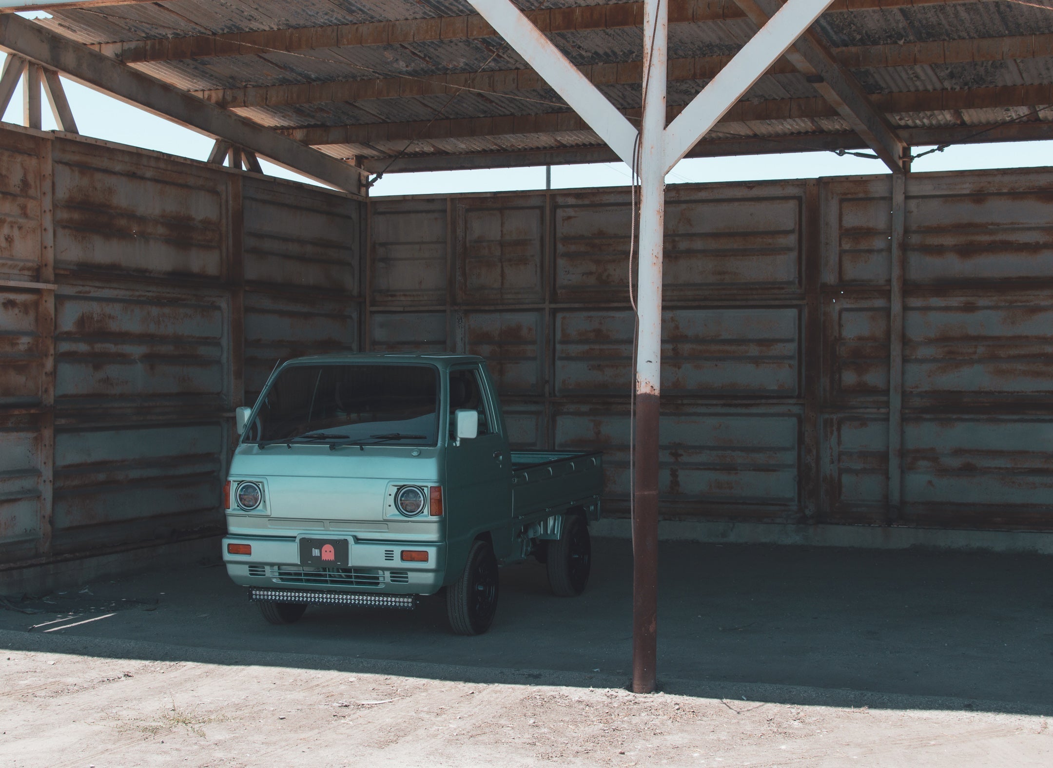 Green Honda Acty kei truck parked beneath a weathered wooden shed, highlighting its compact chassis and sturdy unibody construction ideal for light-duty hauling.