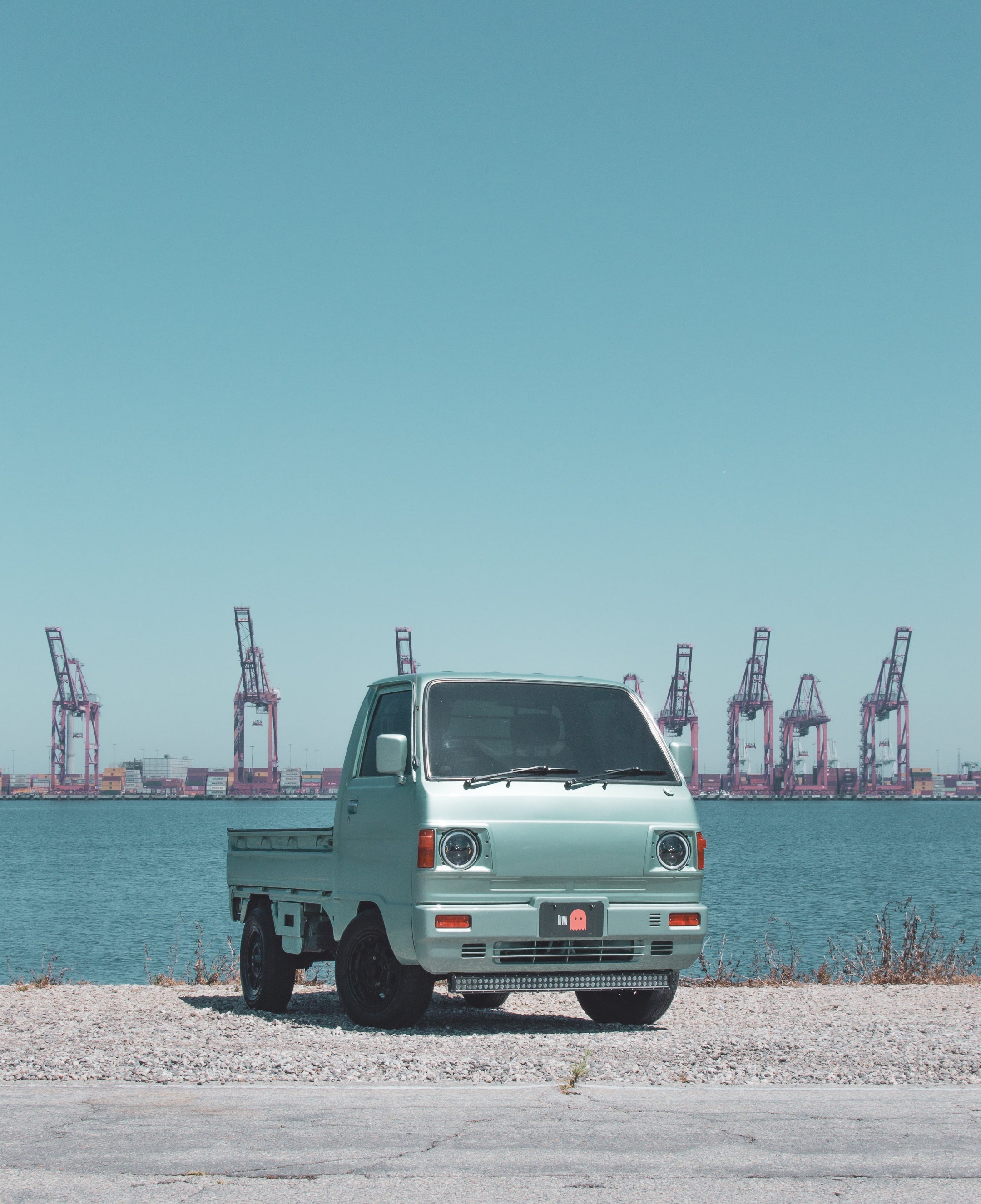 White Suzuki Carry mini truck in motion on urban street, showcasing compact design and fuel efficiency for city transport.
