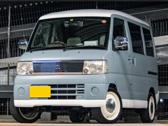 Light blue U62W Mitsubishi Minicab Japanese Kei van with retro white wheels, shown from a front-angle in an urban setting”
