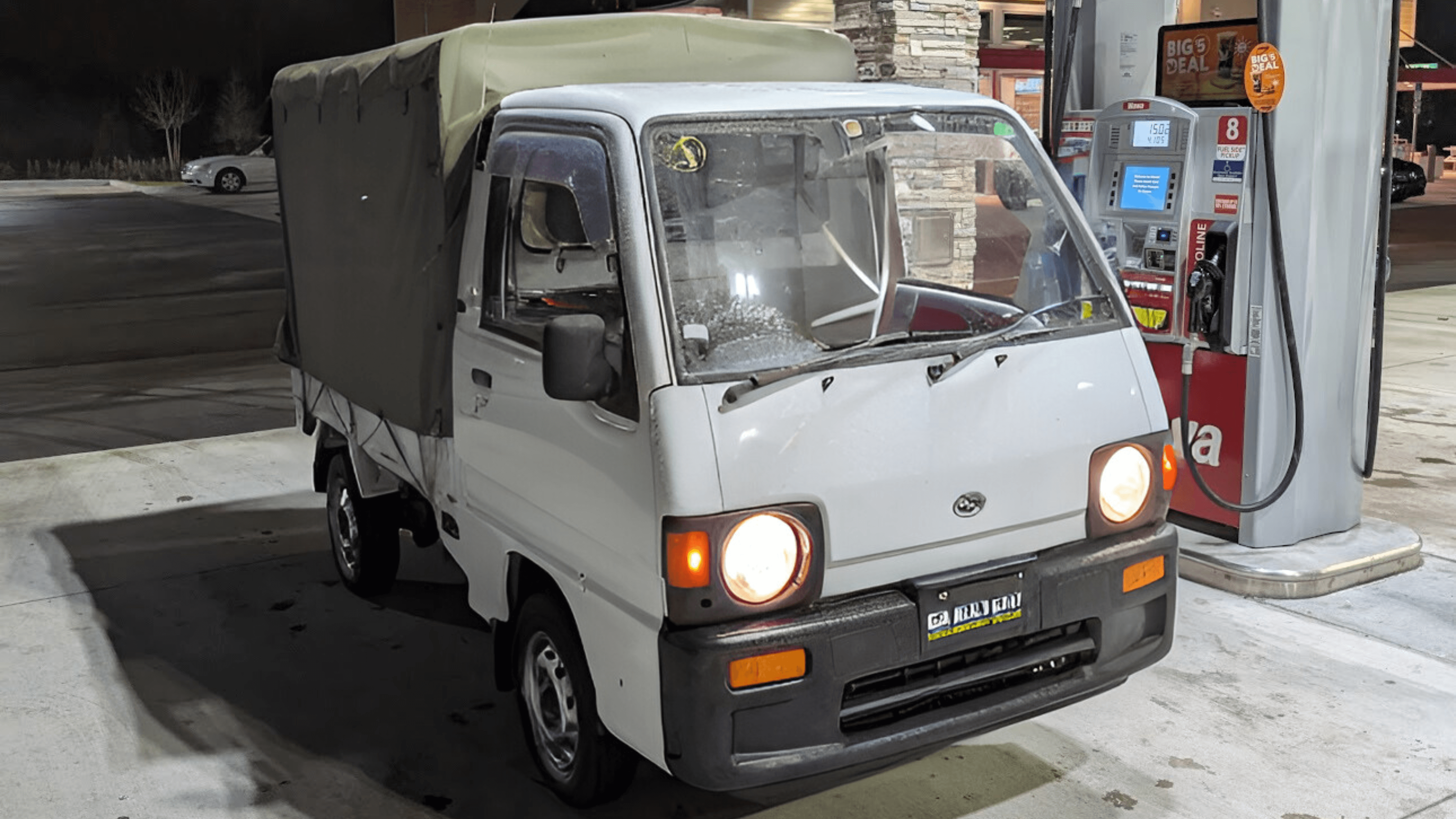 1990 Subaru Sambar KS4 white kei truck with green canvas canopy parked at gas station at night, headlights on, front three quarter view