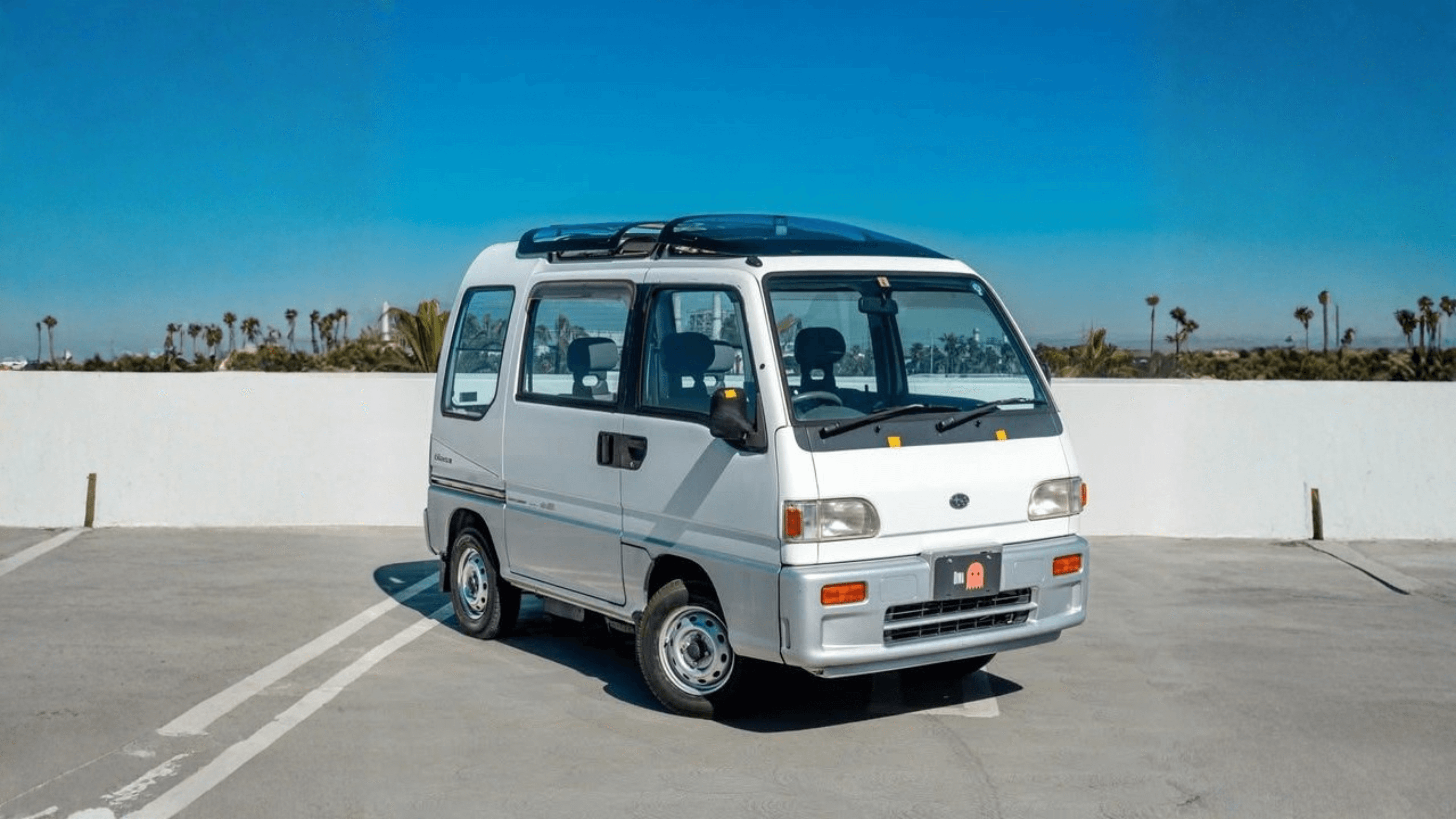 1994 Subaru Sambar KV4 supercharged silver kei van with glass sunroof and roof rack parked on rooftop parking lot under clear blue sky front three quarter view