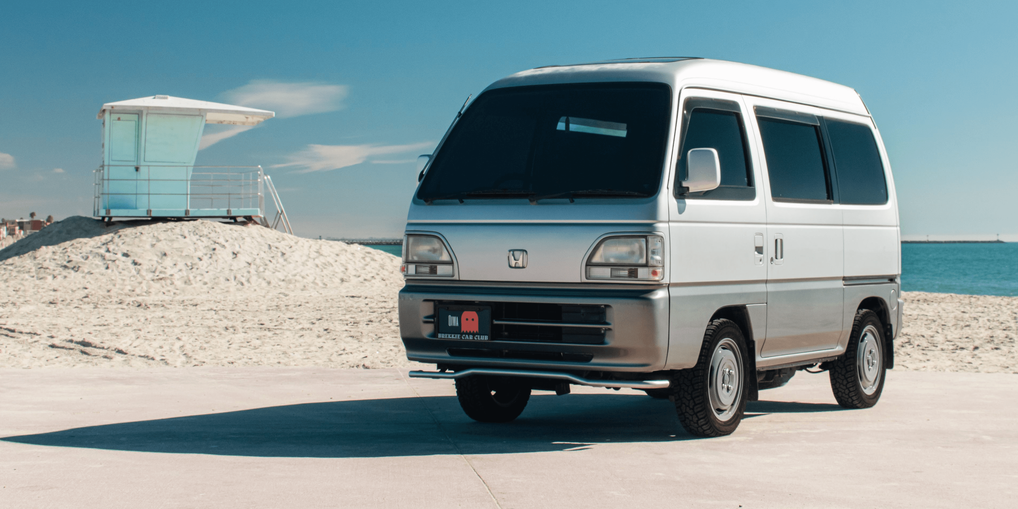 1995 Honda Acty Street HH4 silver kei van parked on sunny beachside pavement near lifeguard tower, front three quarter view showcasing Japanese microvan styling