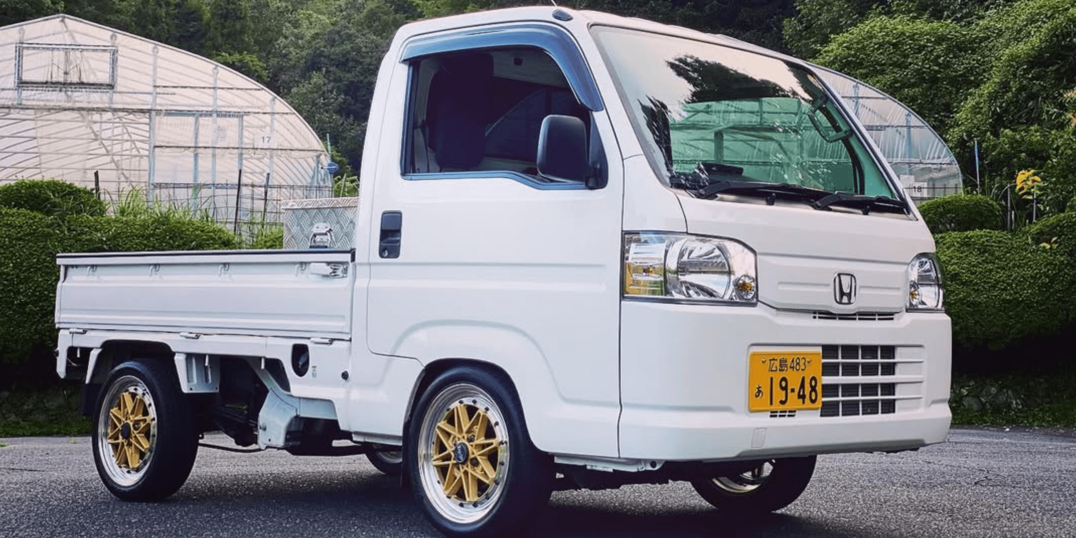 2010 Honda Acty HA9 white kei truck with gold aftermarket wheels parked on rural Japanese farm road in front of greenhouses, low stance front three quarter view