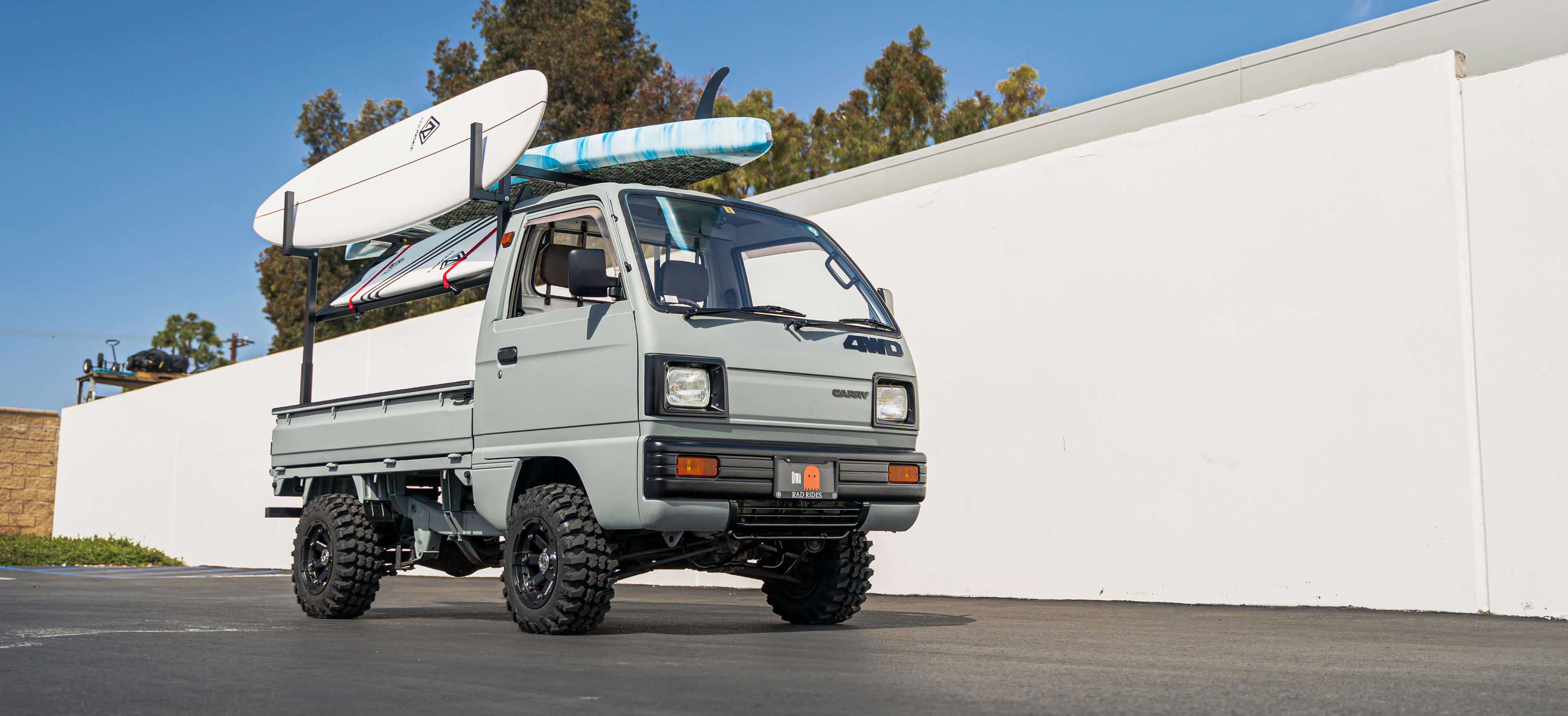 Rugged Suzuki Carry truck modified for off-road use, painted in sleek gray, equipped with oversized tires and carrying multiple surfboards on its rack, parked against a white wall, perfect for adventure sports enthusiasts seeking versatility and performance.