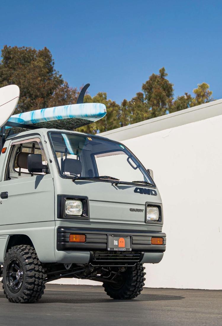 Rugged Suzuki Carry truck modified for off-road use, painted in sleek gray, equipped with oversized tires and carrying multiple surfboards on its rack, parked against a white wall, perfect for adventure sports enthusiasts seeking versatility and performance.