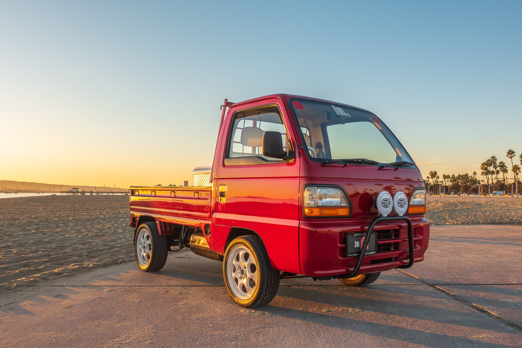 Red Honda Acty HA4 1994 kei truck  with chrome wheels and front bumper guard parked on a beach at sunset with palm trees in the background.