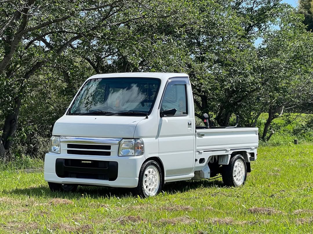 Clean white 2002 Honda Acty HA7 mini truck parked on grass with lush greenery backdrop, highlighting quality Honda Acty parts from Oiwa Garage.