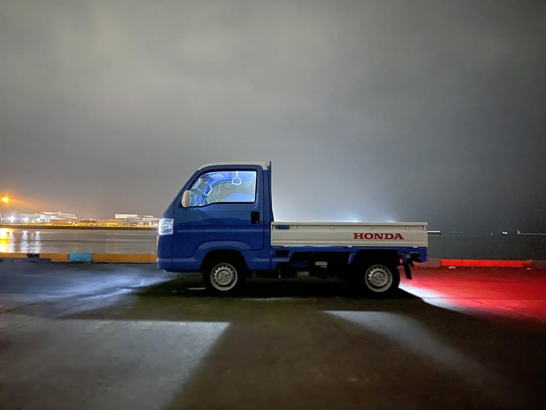 Side profile of a blue and white 2014 Honda Acty Truck HA9 illuminated at night, parked along a waterfront with city lights reflecting off the water, emphasizing its compact build and striking two-tone color scheme.