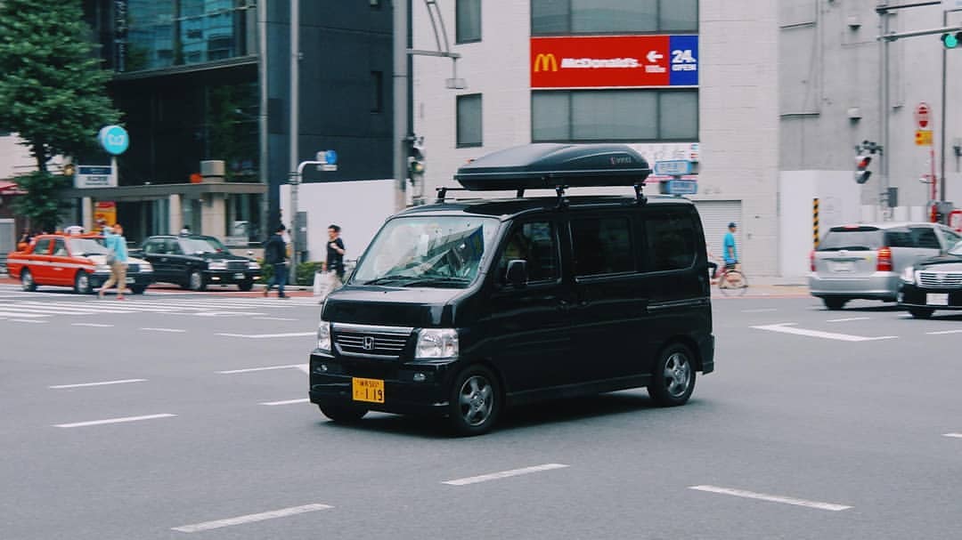 ront-side view of a black 2011 Honda Vamos HM2 driving in a busy urban intersection, equipped with a rooftop cargo carrier, highlighting its versatility as a compact city van.