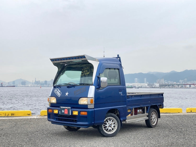 Blue 1995 Subaru Sambar KS4 kei truck with chrome sun‑visor and alloy wheels parked on a waterfront pier, Japanese city skyline and distant mountains under an overcast sky.