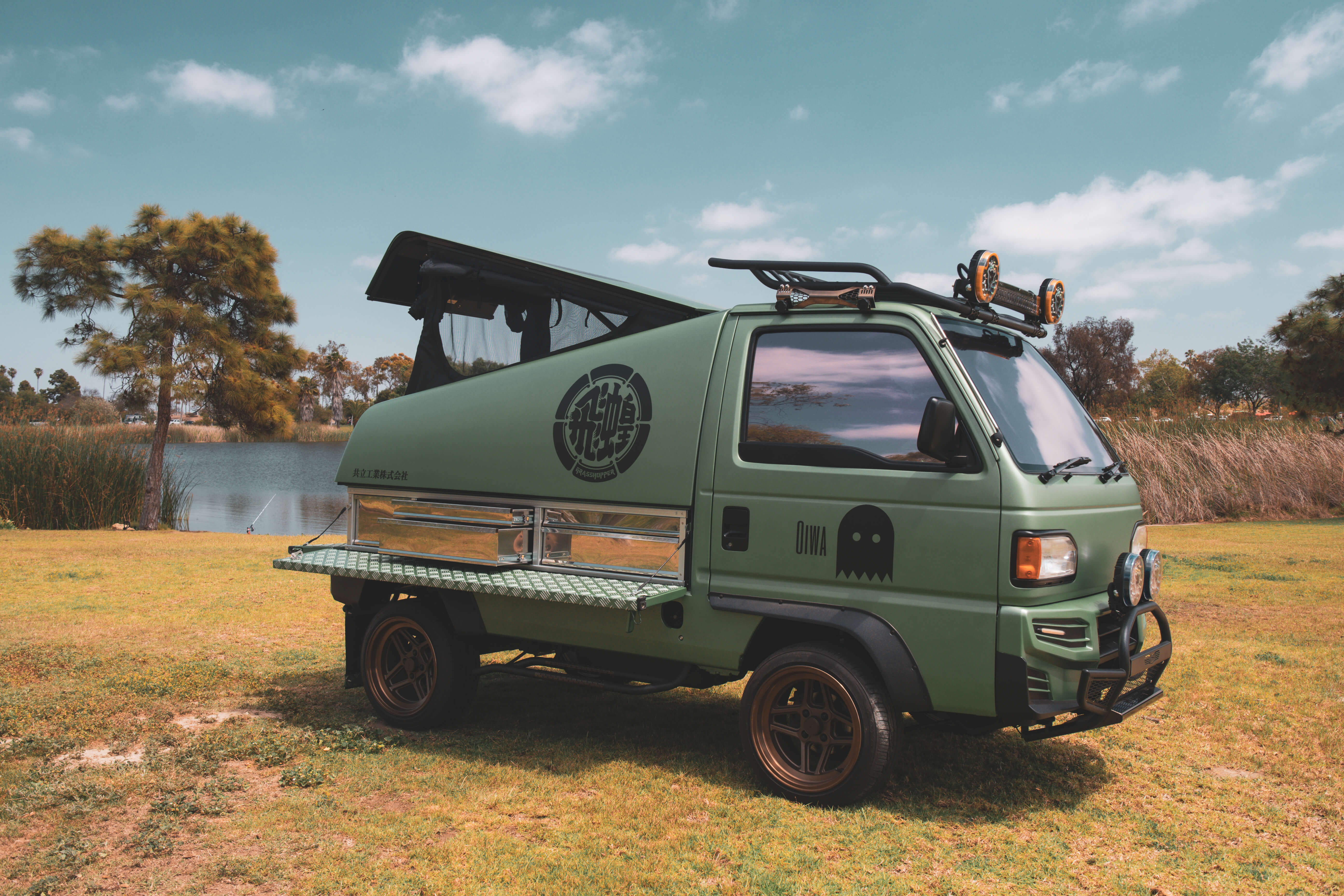 Customized Honda Acty mini truck by Oiwa Garage set in an outdoor landscape, featuring off-road upgrades, Grasshopper camper shell, and rugged aesthetics for adventure.