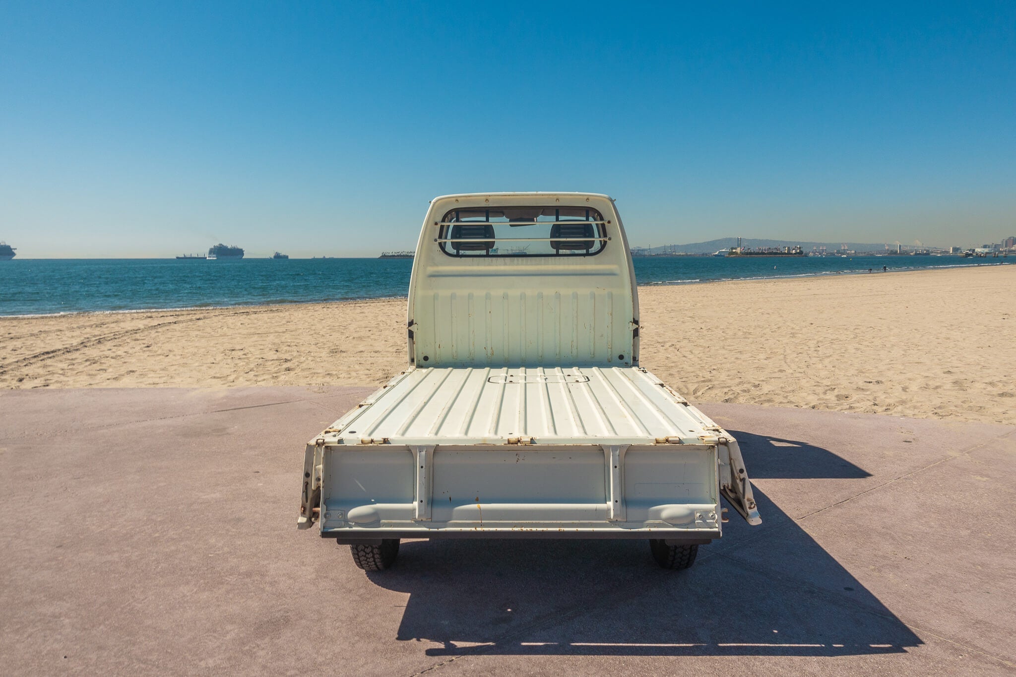 Rear view of a Honda Acty truck bed facing a sandy beach and ocean under a clear blue sky.