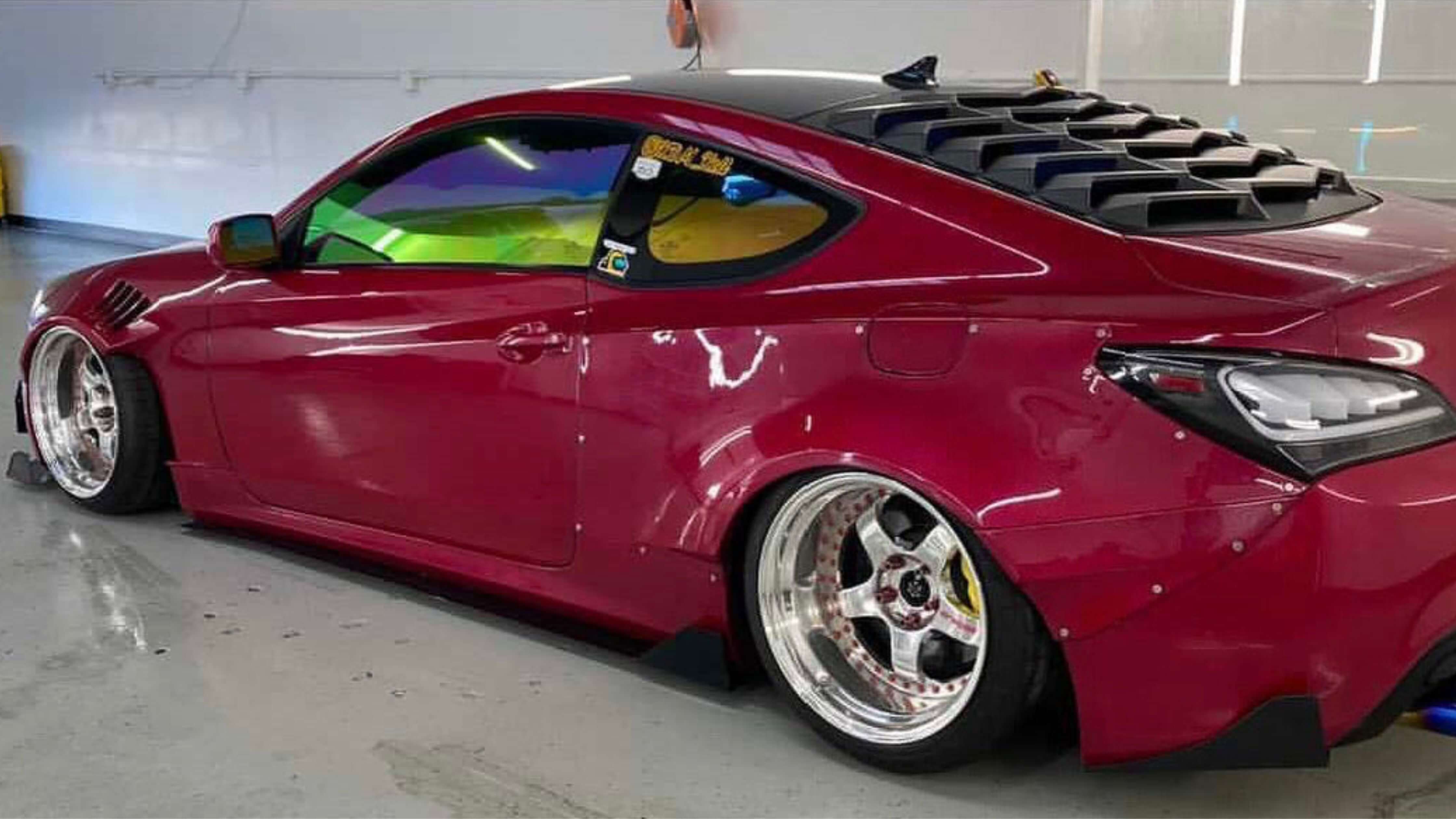 A modified red sports car with wide fenders, custom wheels, and a louvered rear window parked indoors after tinting.