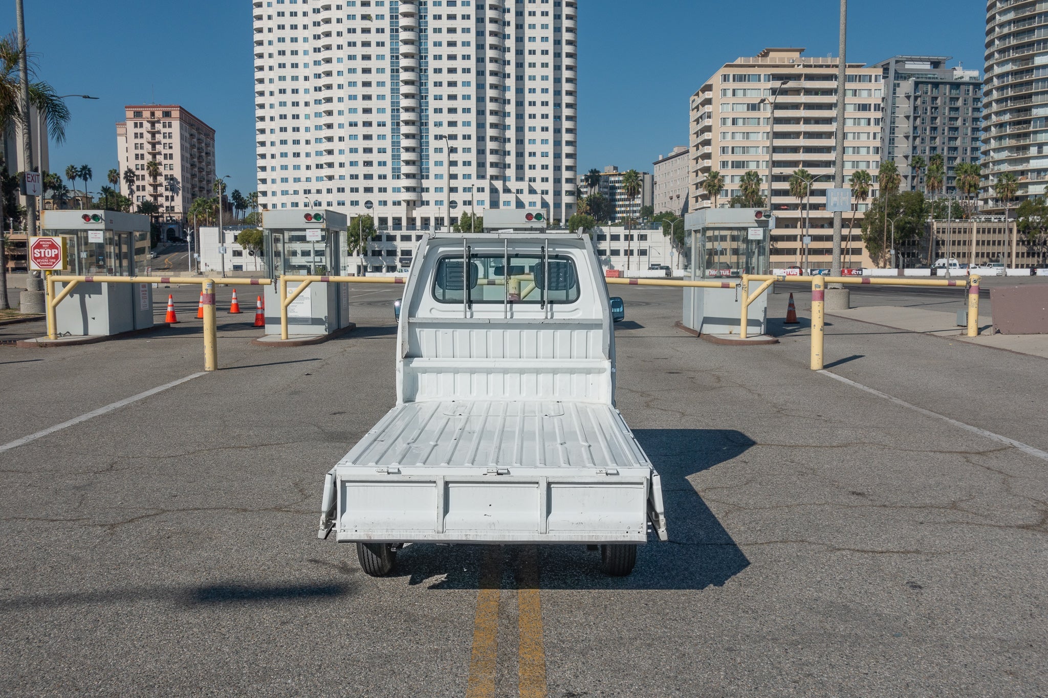 A small white flatbed truck is parked in front of a closed gate at a parking lot, with tall buildings in the background.