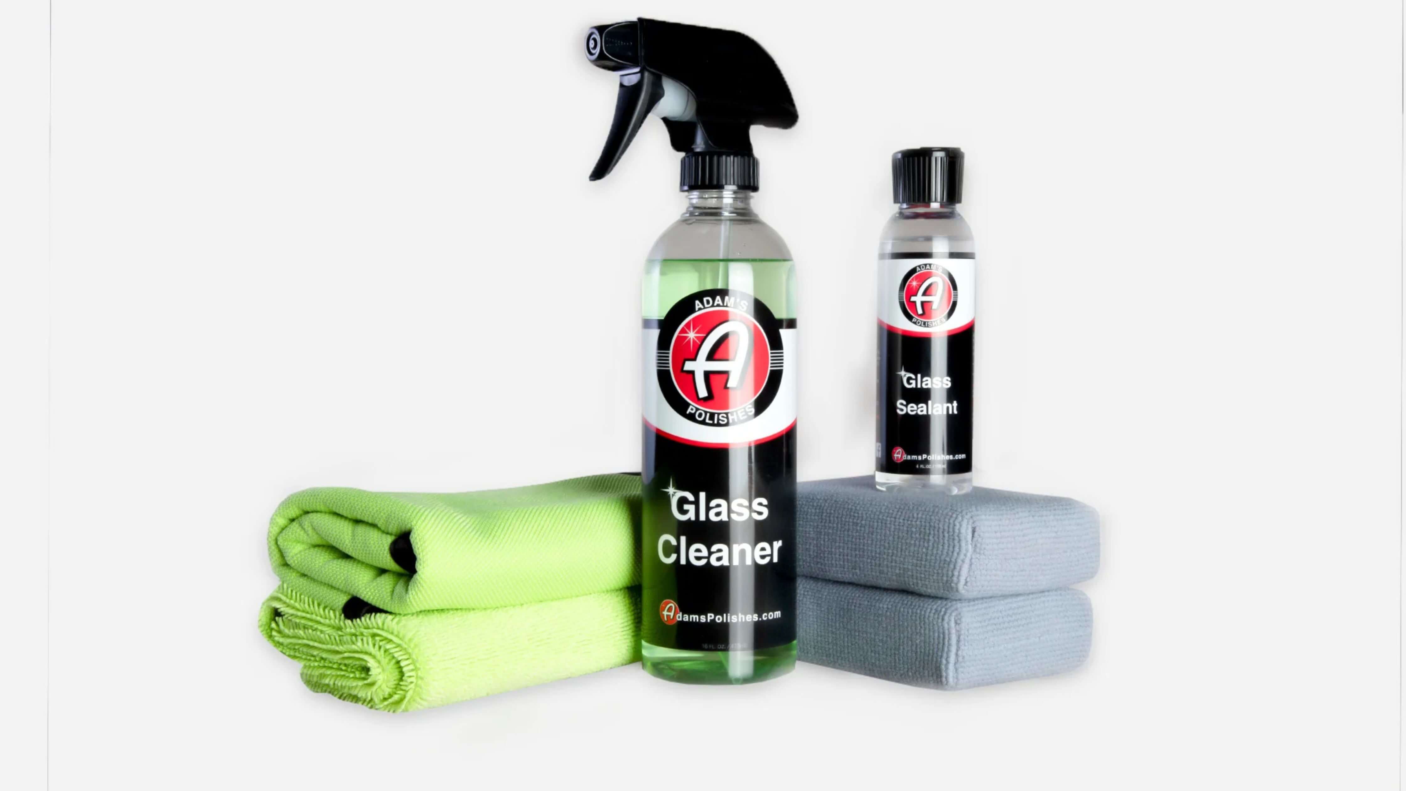 Auto glass cleaning products including a spray bottle of Adam's Polishes cleanser, two microfiber cloths, and sponges for streak-free shine.