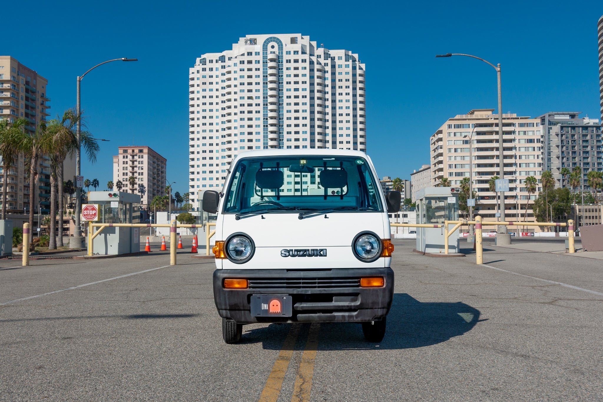 A white Suzuki van is parked on a street with tall buildings and palm trees in the background.