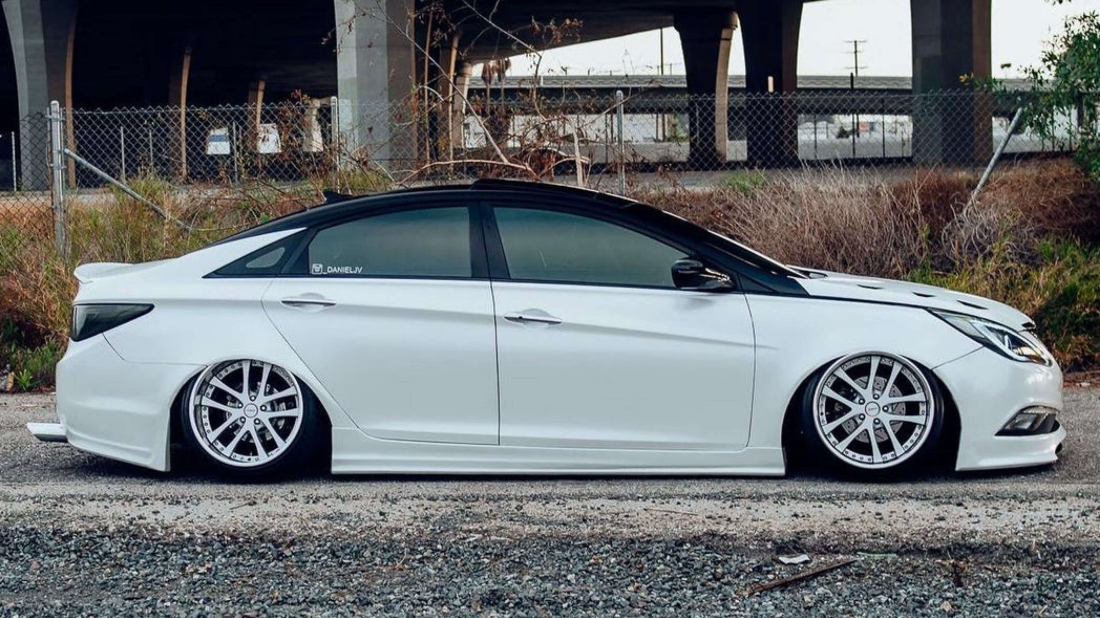 A white car with tinted windows parked on a gravel surface near a chain-link fence and overpass.