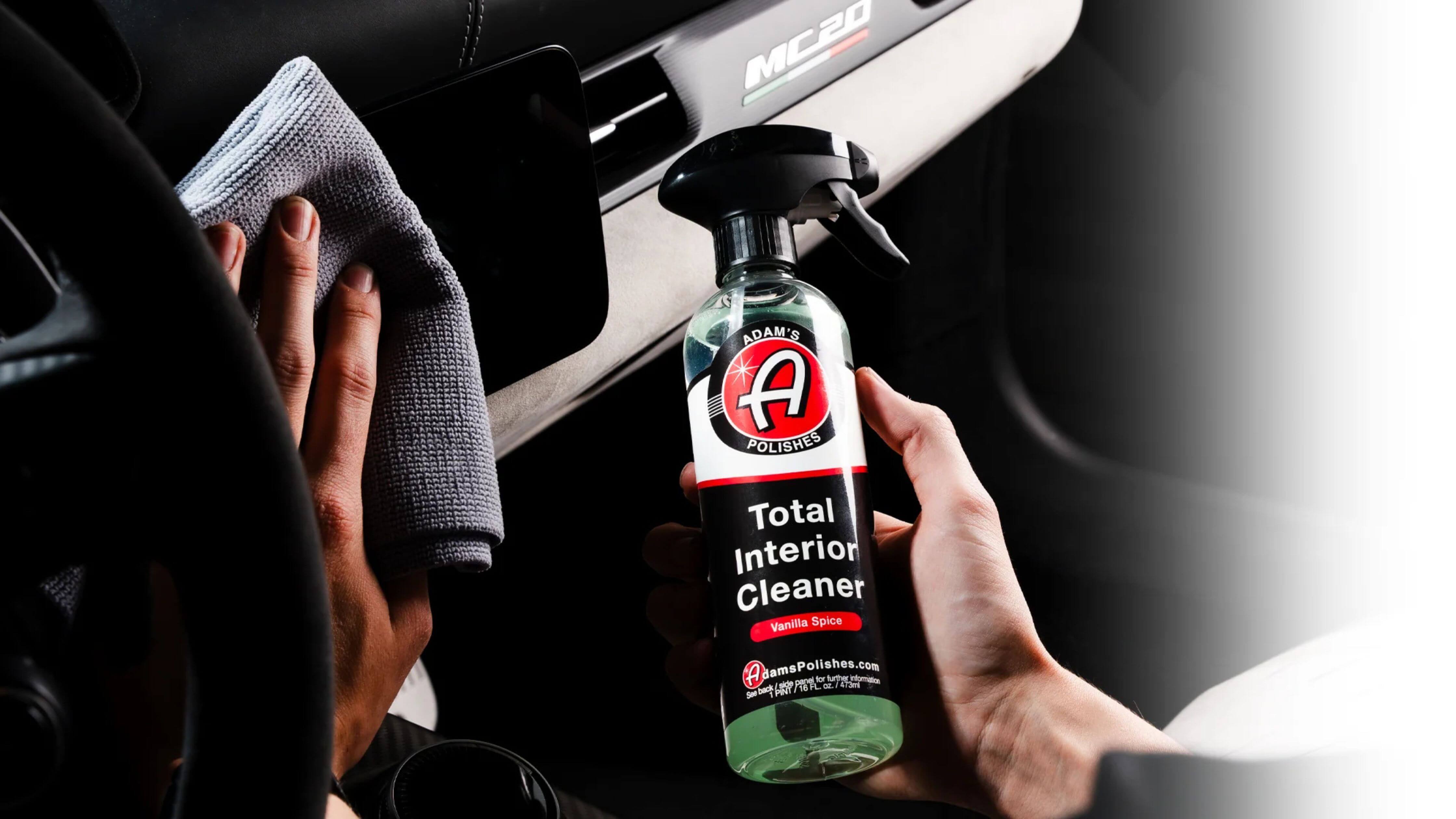 A person is cleaning a car interior with a microfiber cloth and holding a bottle of Adam's Polishes Total Interior Cleaner.