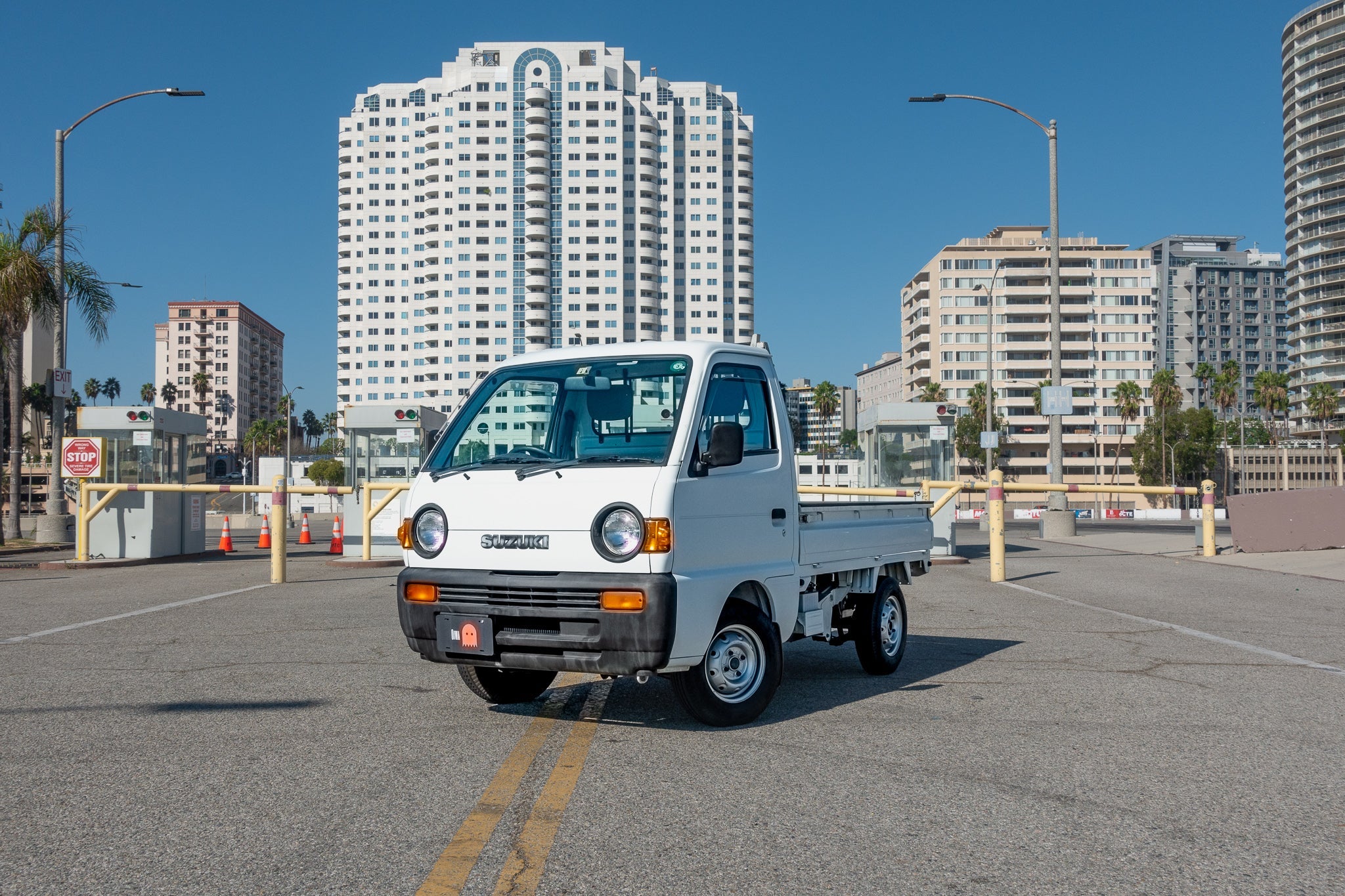 A white Suzuki mini truck is parked on a city street with tall buildings in the background.