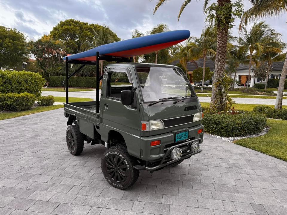 Customized Subaru Sambar with military green paint, roof rack holding a kayak, upgraded black off-road tires, and front-mounted spotlights parked in a tropical driveway.