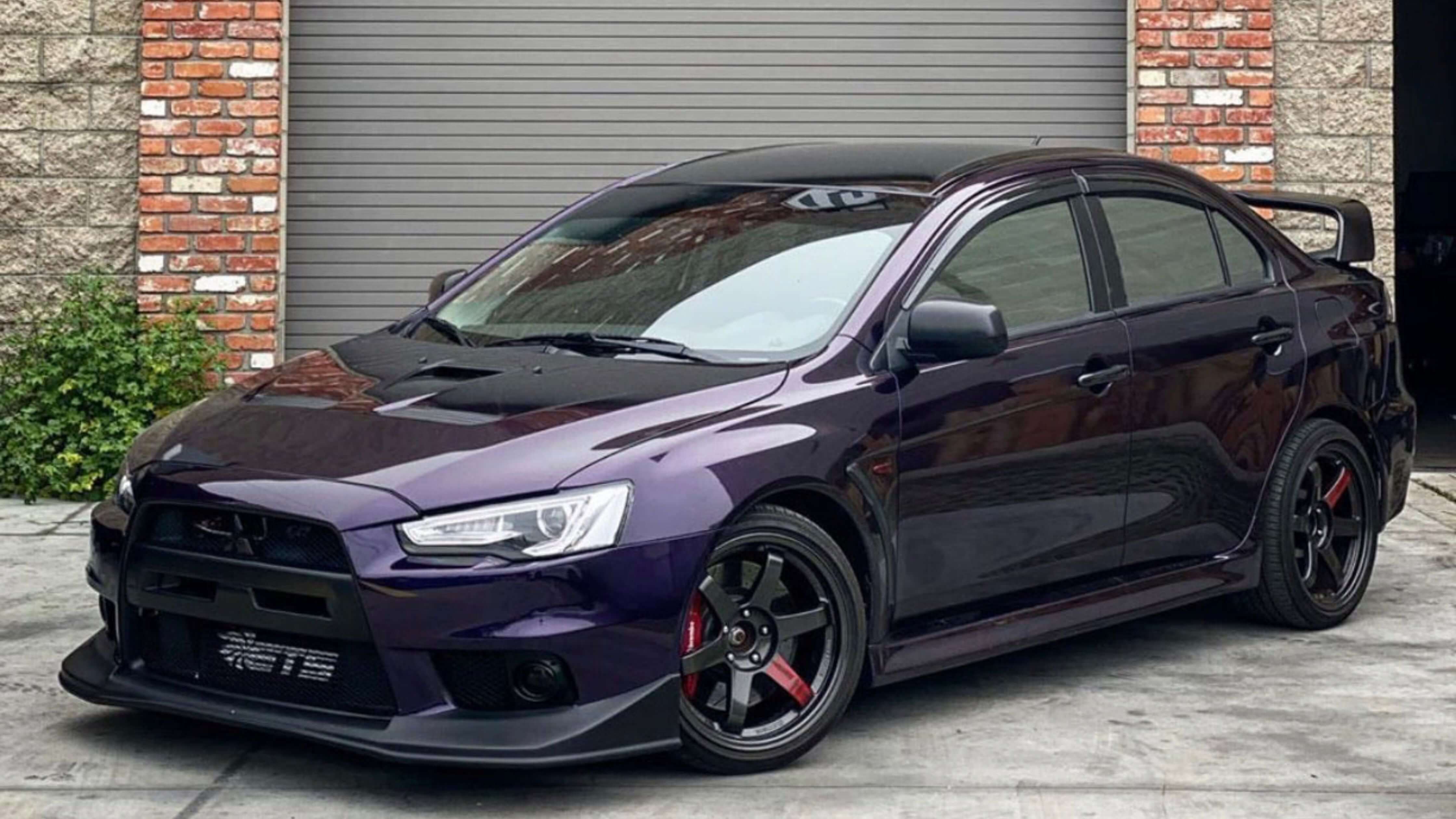 A dark purple sports sedan with black rims and a rear spoiler parked in front of a brick and stone building.