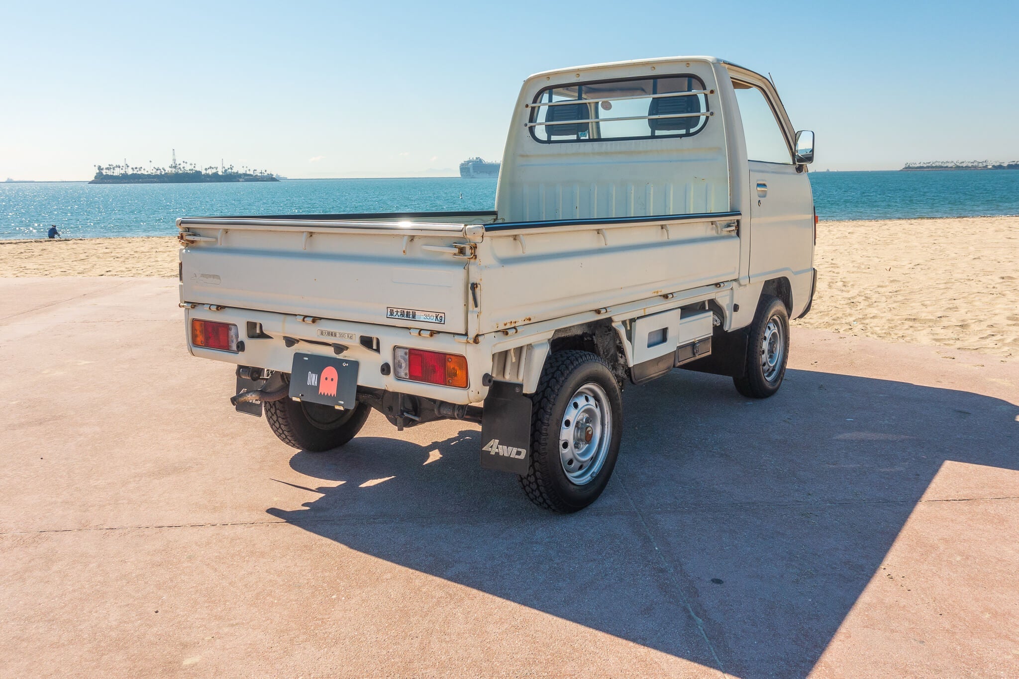 Honda Acty mini truck parked on a beachside pavement with ocean and island in the background.