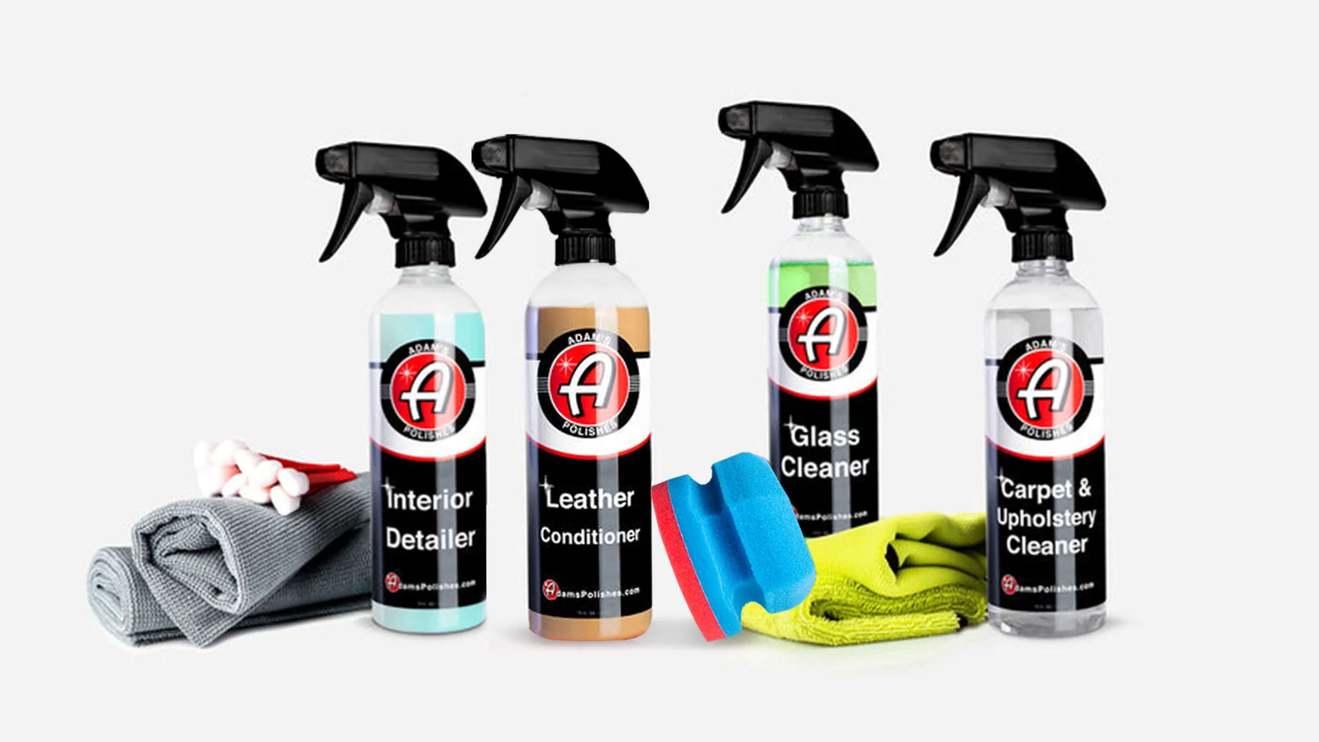 Four spray bottles of car cleaning products labeled 'Interior Detailer,' 'Leather Conditioner,' 'Glass Cleaner,' and 'Carpet & Upholstery Cleaner,' accompanied by cleaning cloths, cotton swabs, and a sponge.