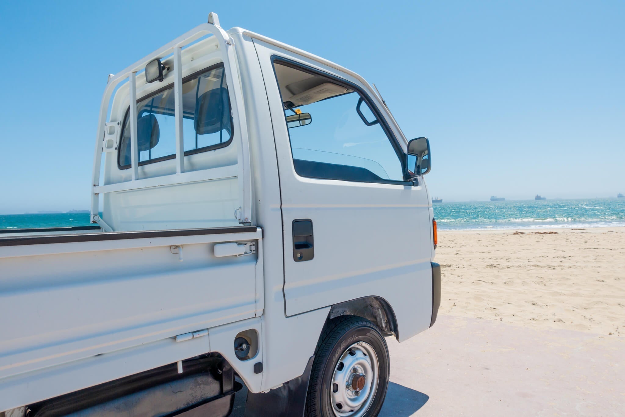 White Honda Acty truck parked on a sunny beach with ocean in the background.