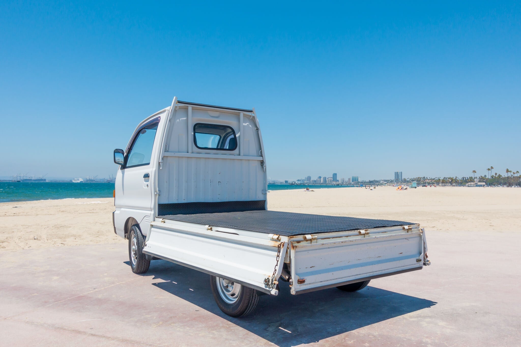 Honda Acty mini truck parked on a sunny beach with city skyline in the background.