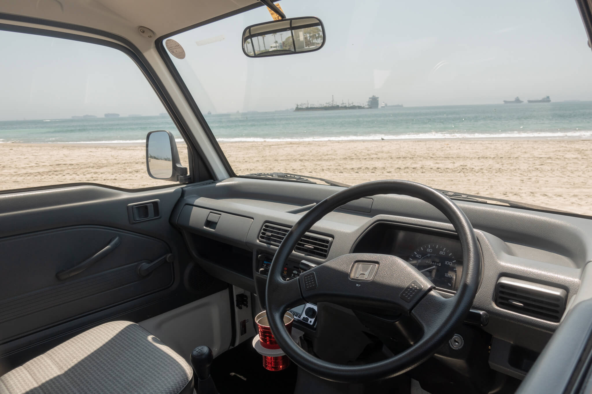 Honda Acty interior view with steering wheel, dashboard, and beach scenery through the window.