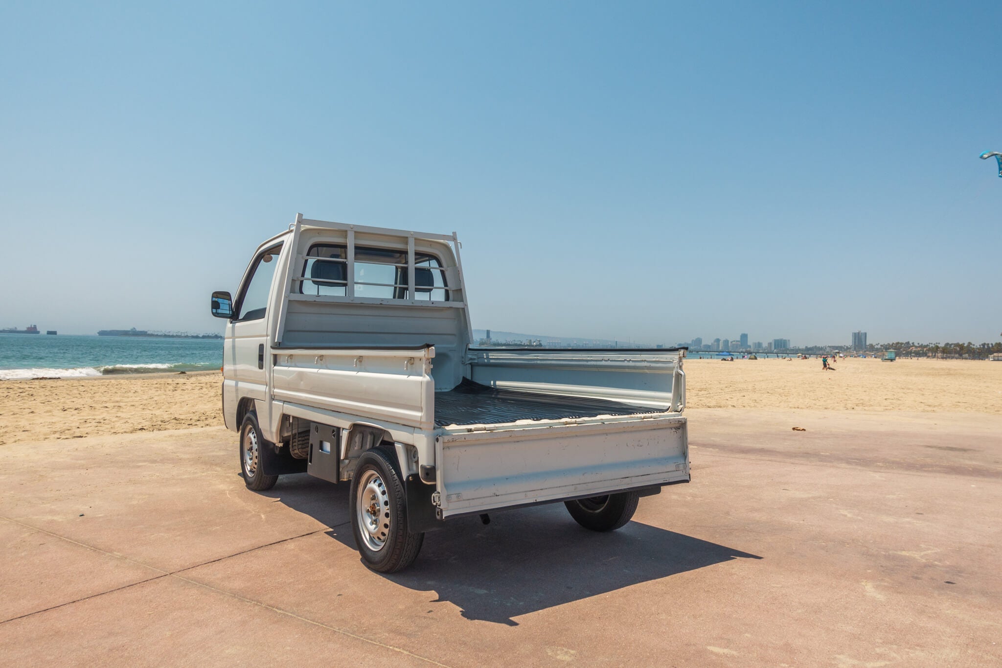 Honda Acty mini truck parked on a beach with ocean and city skyline in the background.