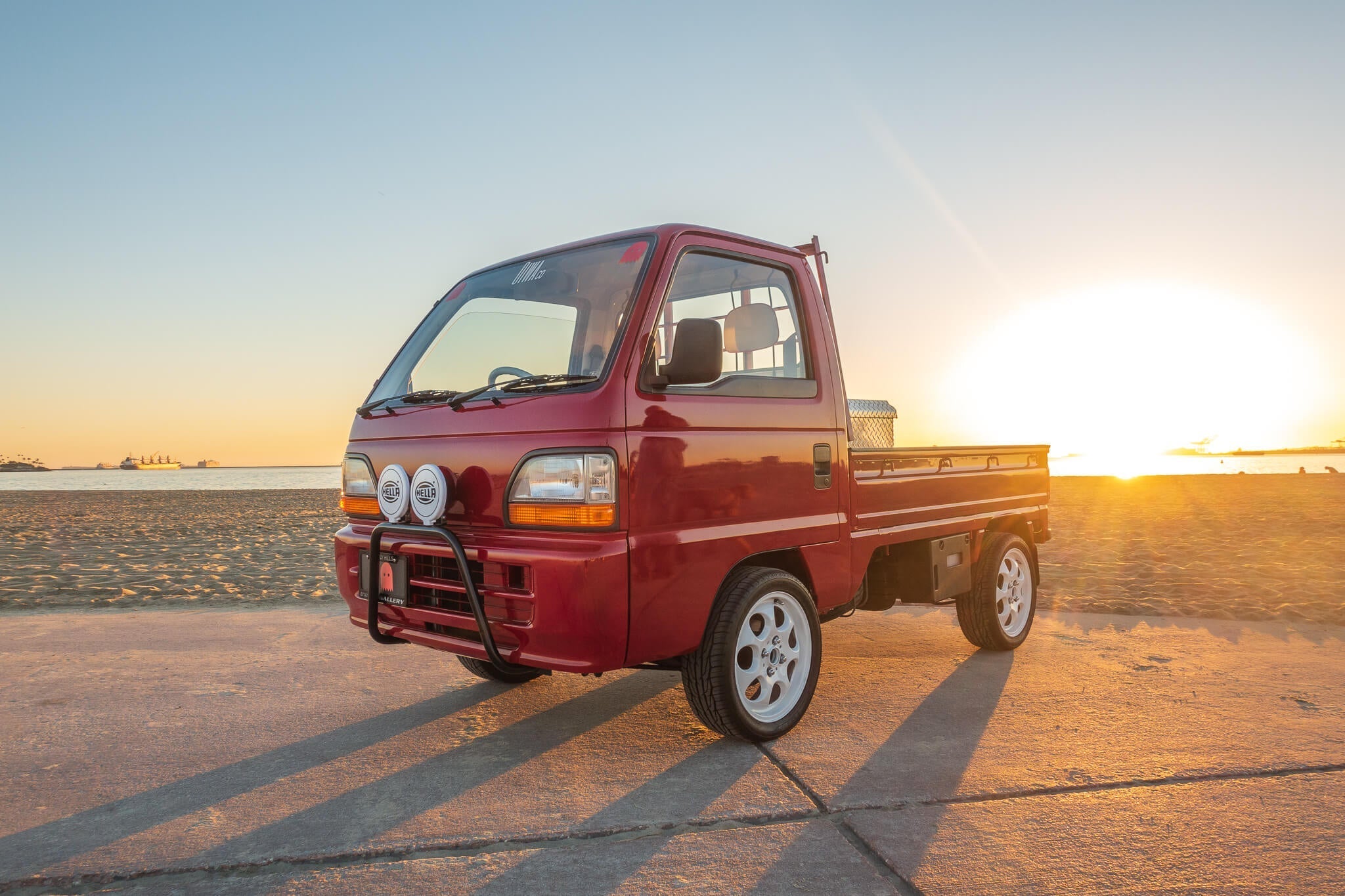 Red 1995 Honda Acty truck featuring aftermarket fog lights, alloy wheels, and bull bar, captured during a sunset on a beachside promenade.