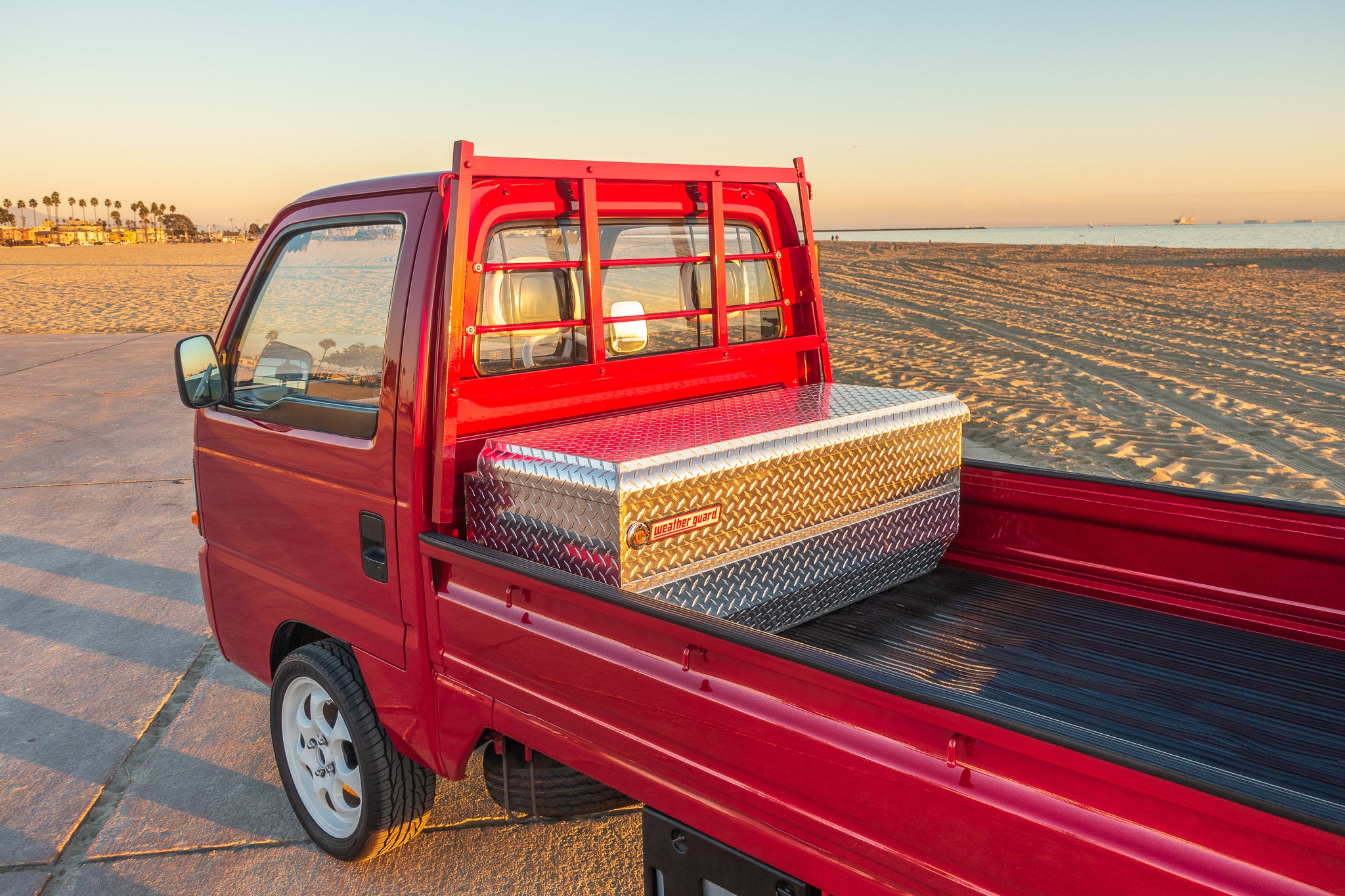 Red Kei truck parked on sandy beach at sunset, showcasing compact design and utility features including a metal toolbox in the bed, ideal for highlighting its versatility and appeal in scenic outdoor settings.